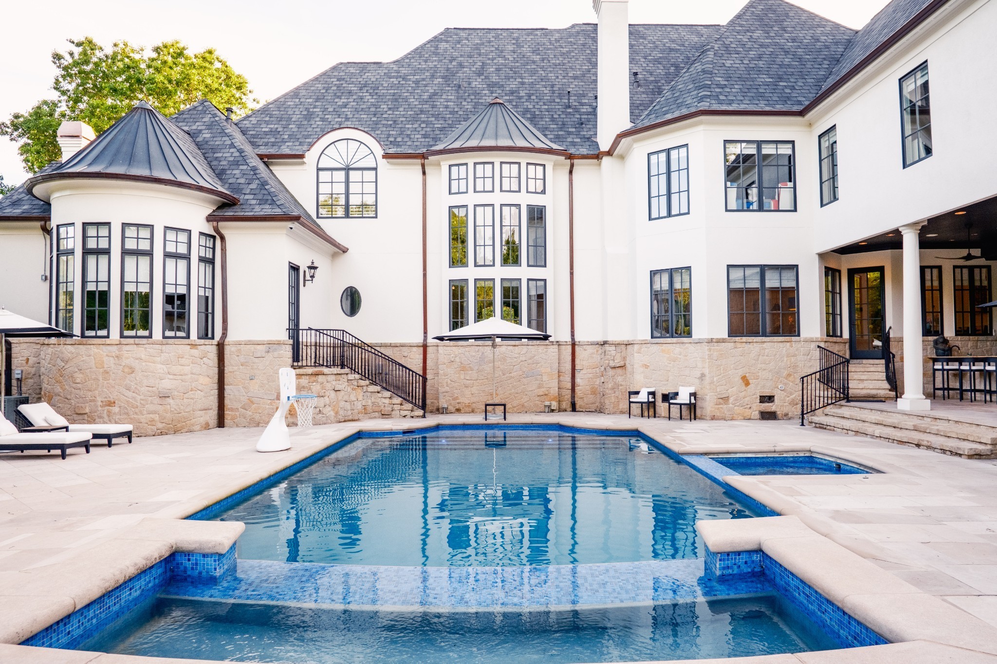 Large white home behind a blue pool and raised spa, with stone walls and a basketball hoop.