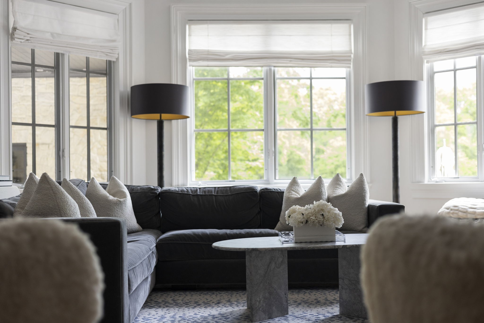 Living room with a dark sectional, marble coffee table, white flowers, and tall windows.