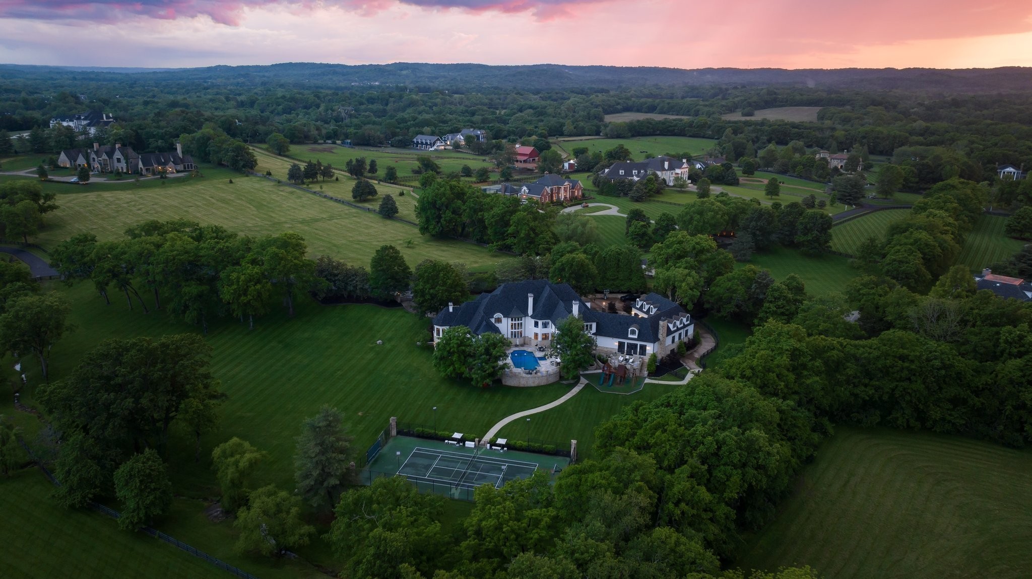 Aerial view of a large estate with a pool, expansive lawns, and tennis court at dusk.