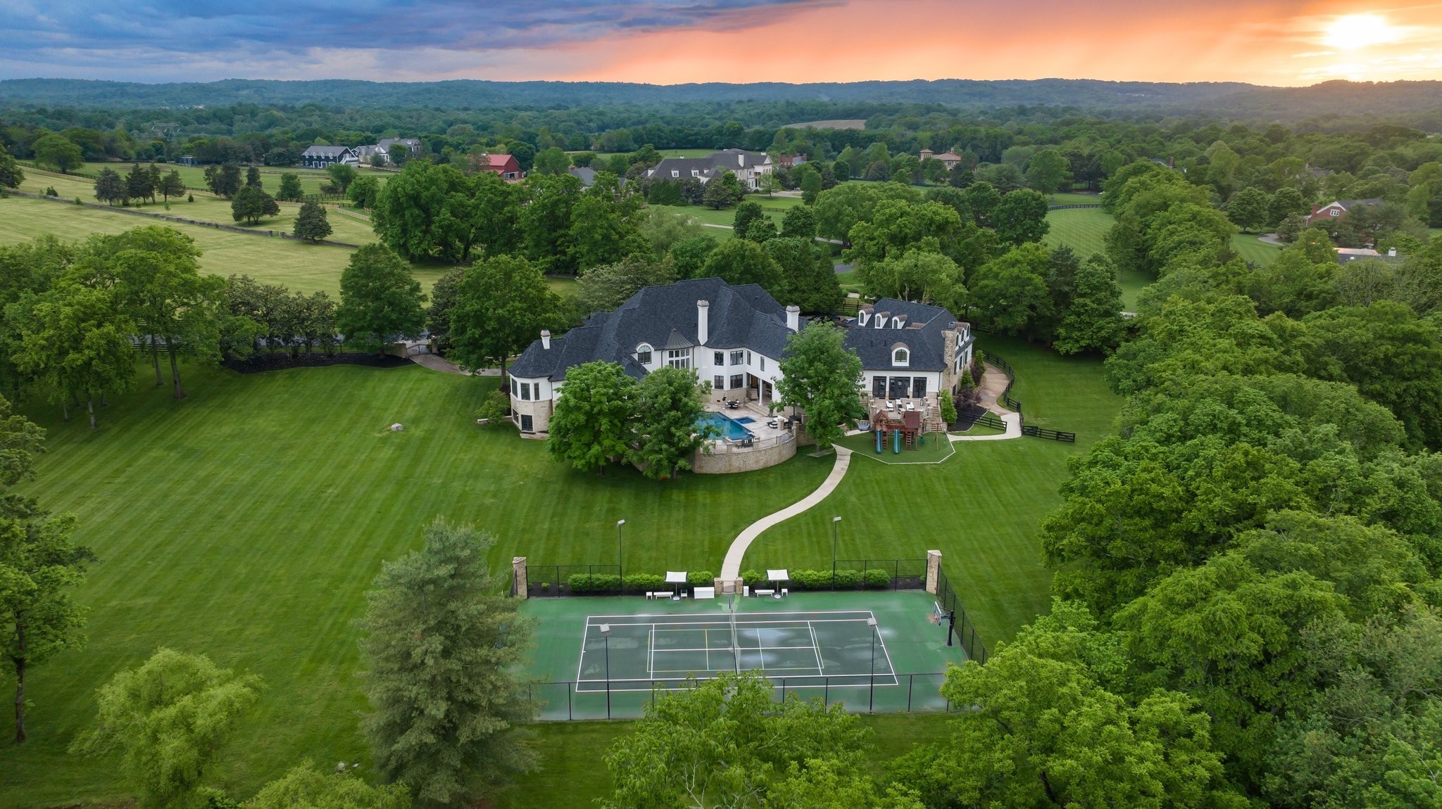 Aerial view of a large estate with a pool, tennis court, and broad lawns at sunset.