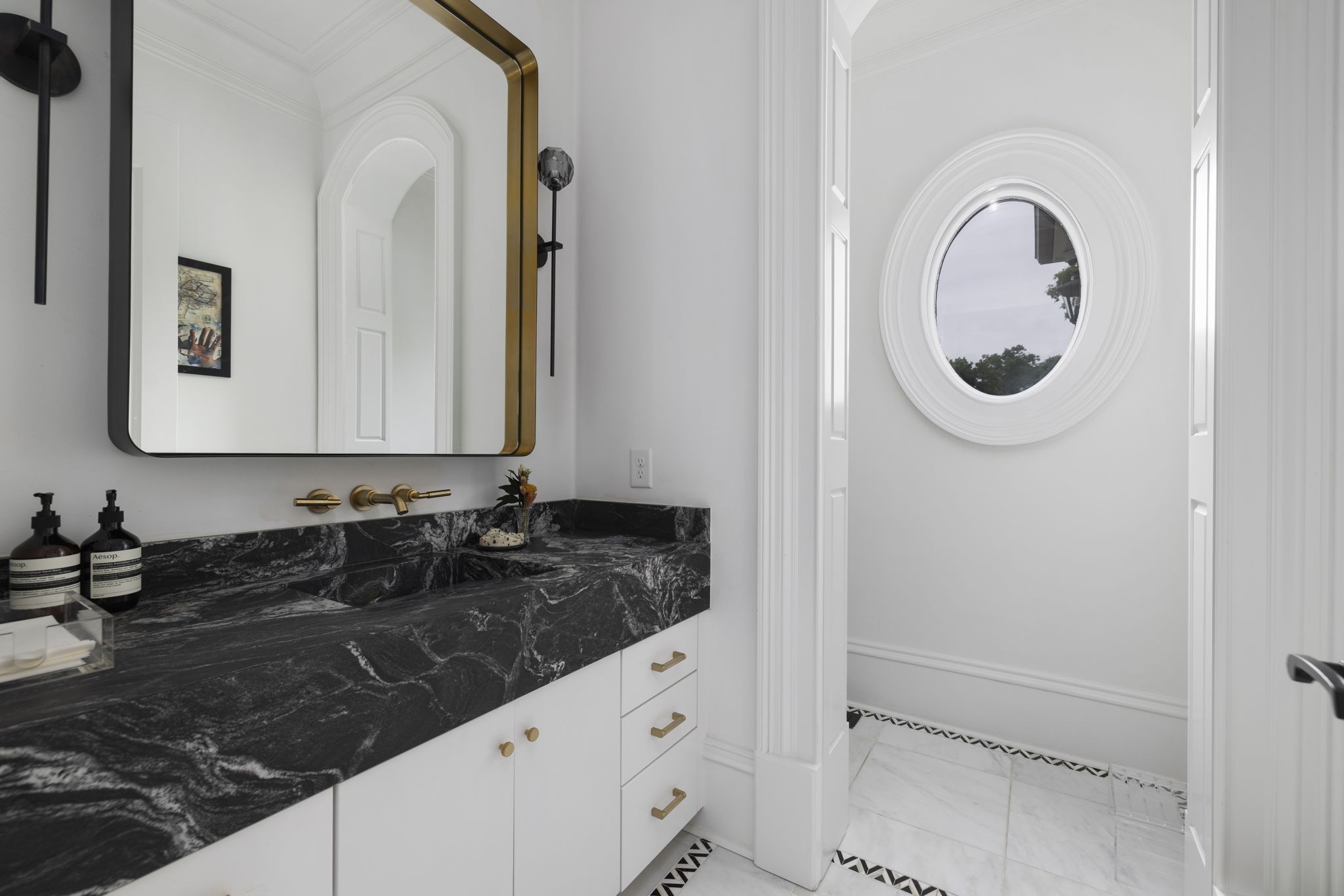 Bathroom vanity with black veined stone, brass fixtures, and an adjoining room with a round window.
