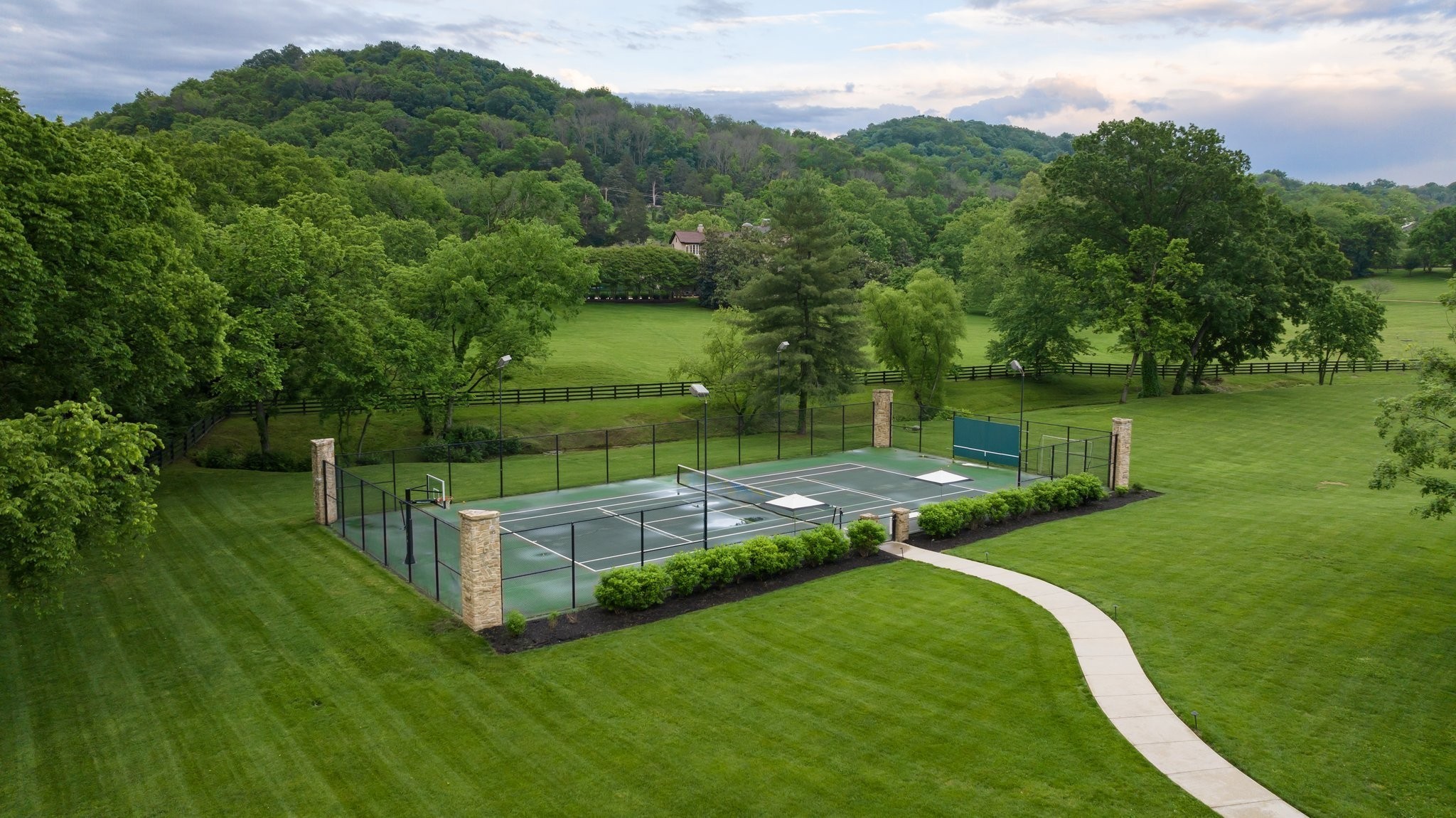 Fenced tennis and basketball court amid manicured lawns, mature trees, and wooded hills.