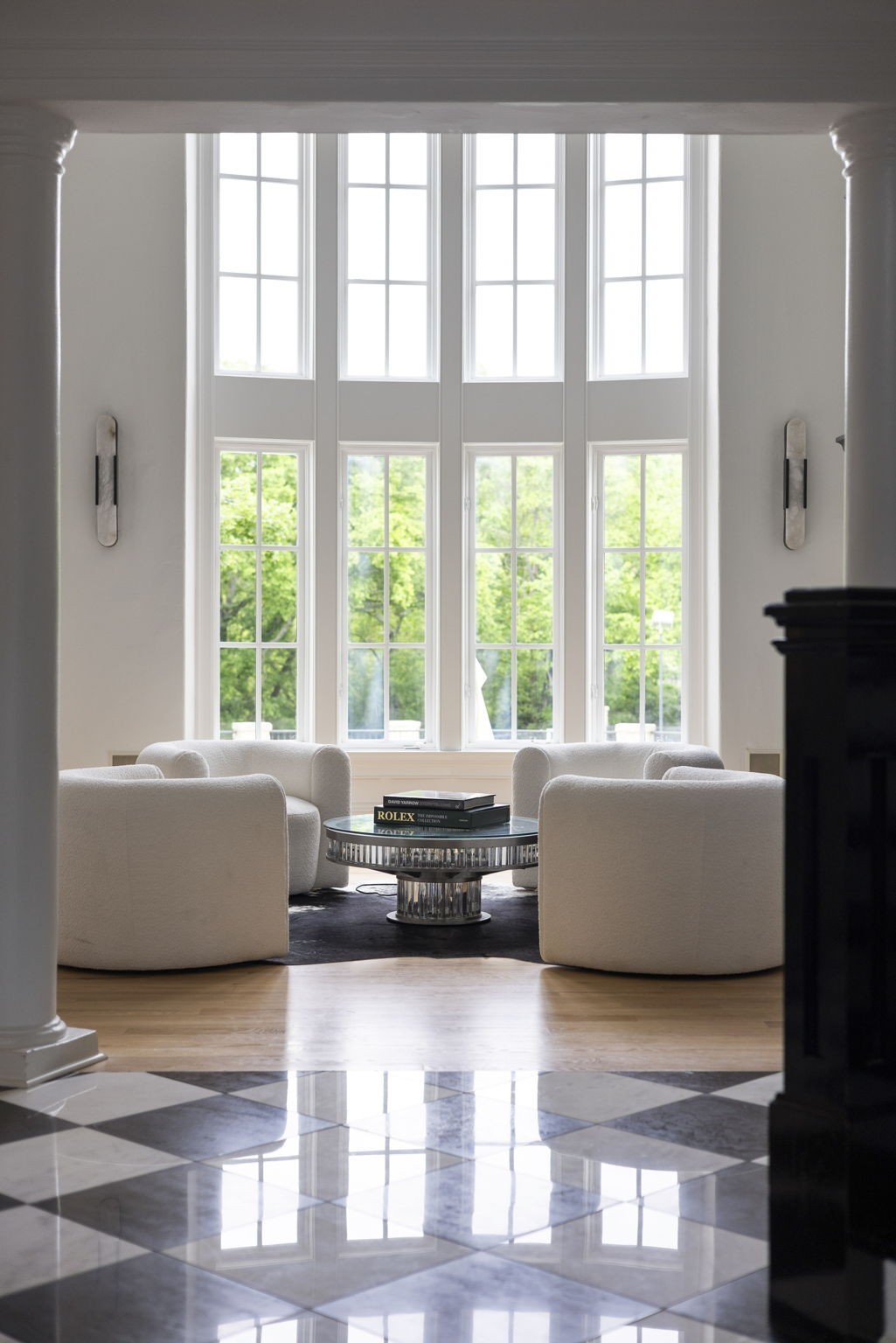 Living room with white chairs, a round glass coffee table, and tall windows overlooking leafy trees.