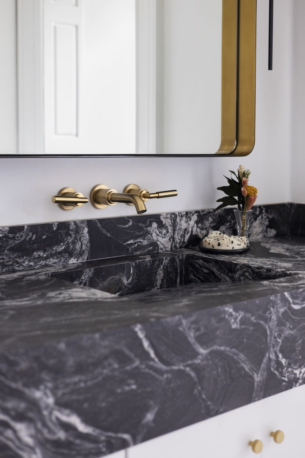 Bathroom vanity with a dark veined stone sink, brass wall-mounted faucet, and gold-framed mirror.