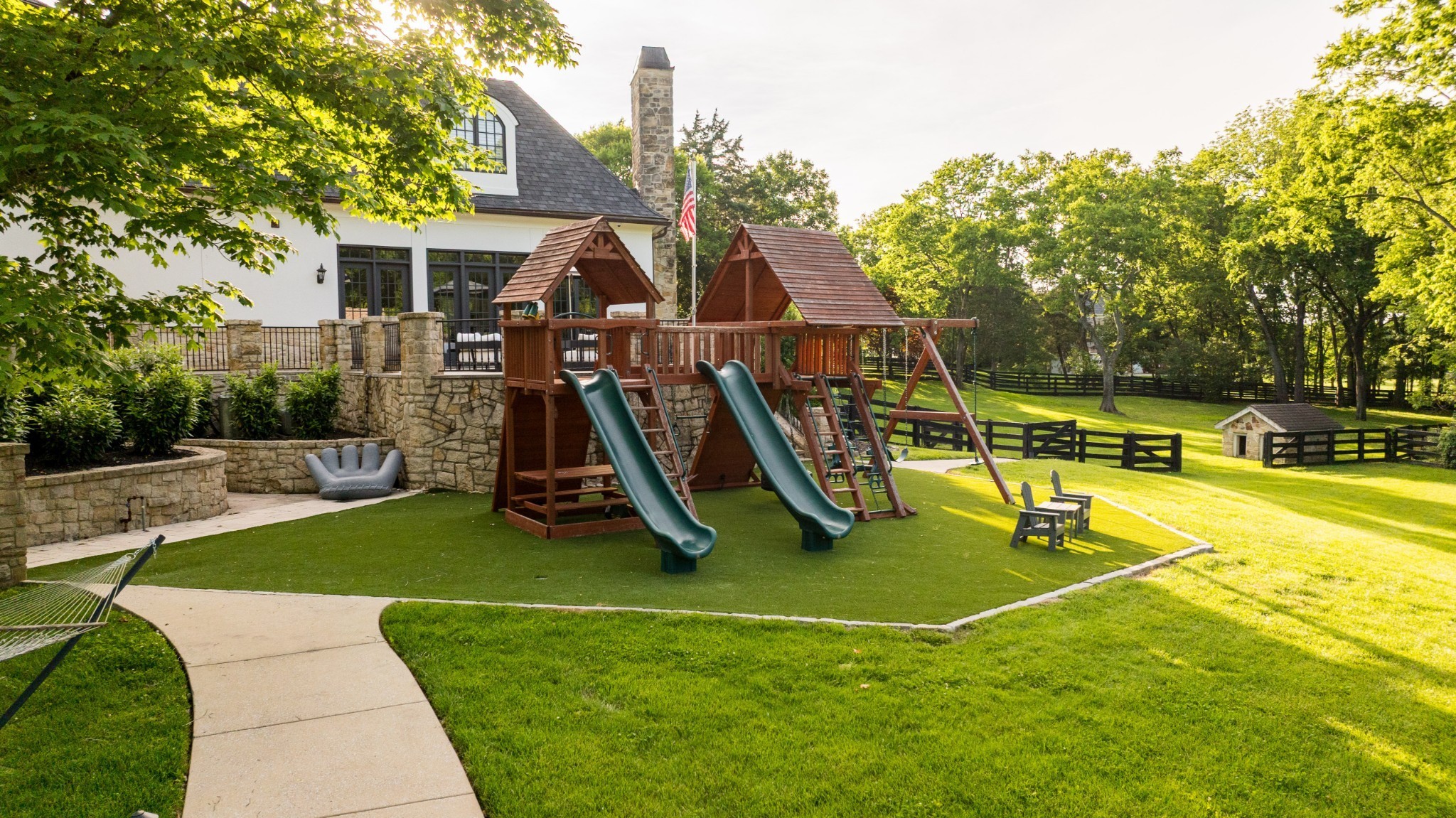 Backyard with a wooden playset, stone patio walls, lawn, and tree-lined fenced grounds.