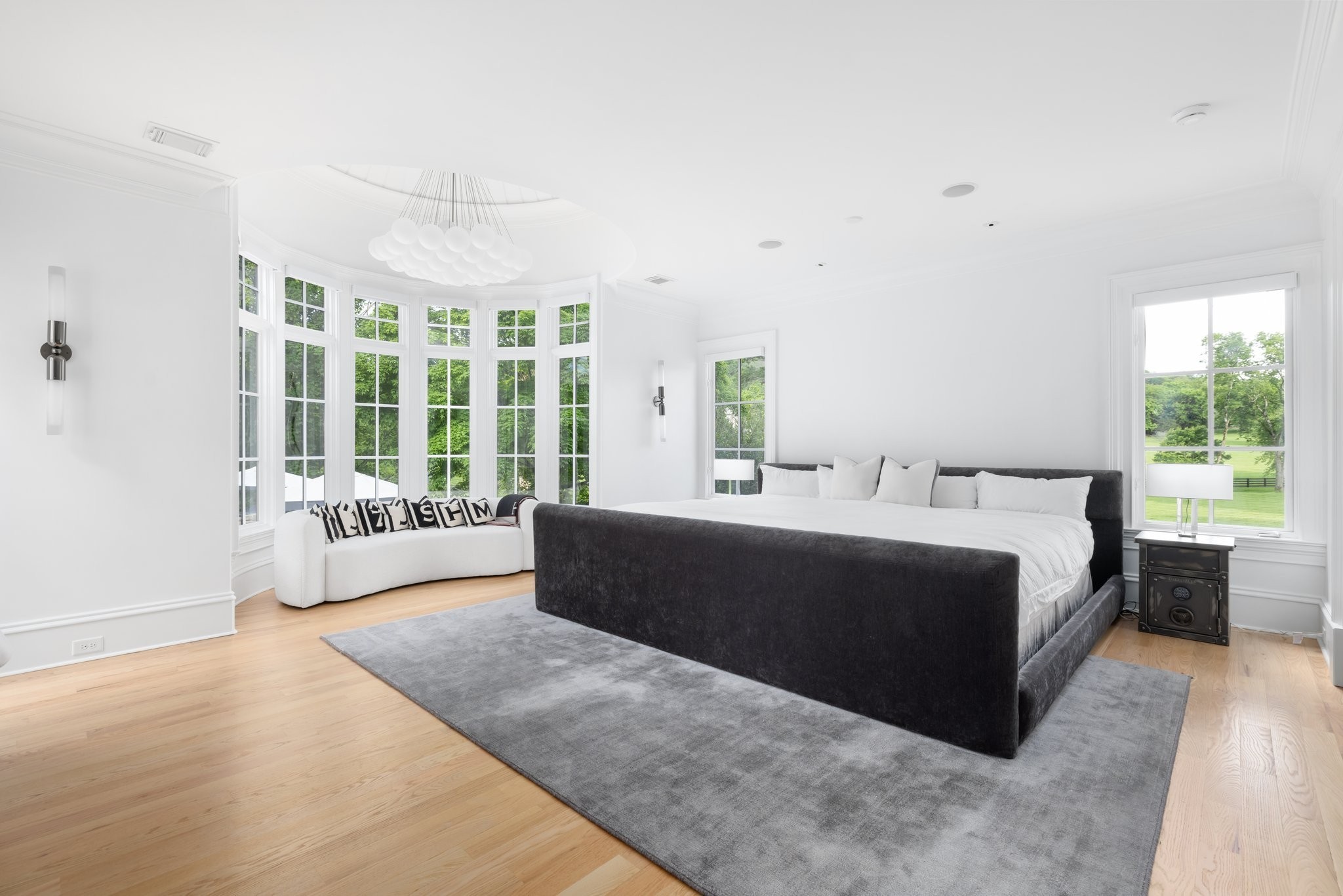 Bright bedroom with a dark upholstered bed, gray rug, white sofa, and curved wall of tall windows.