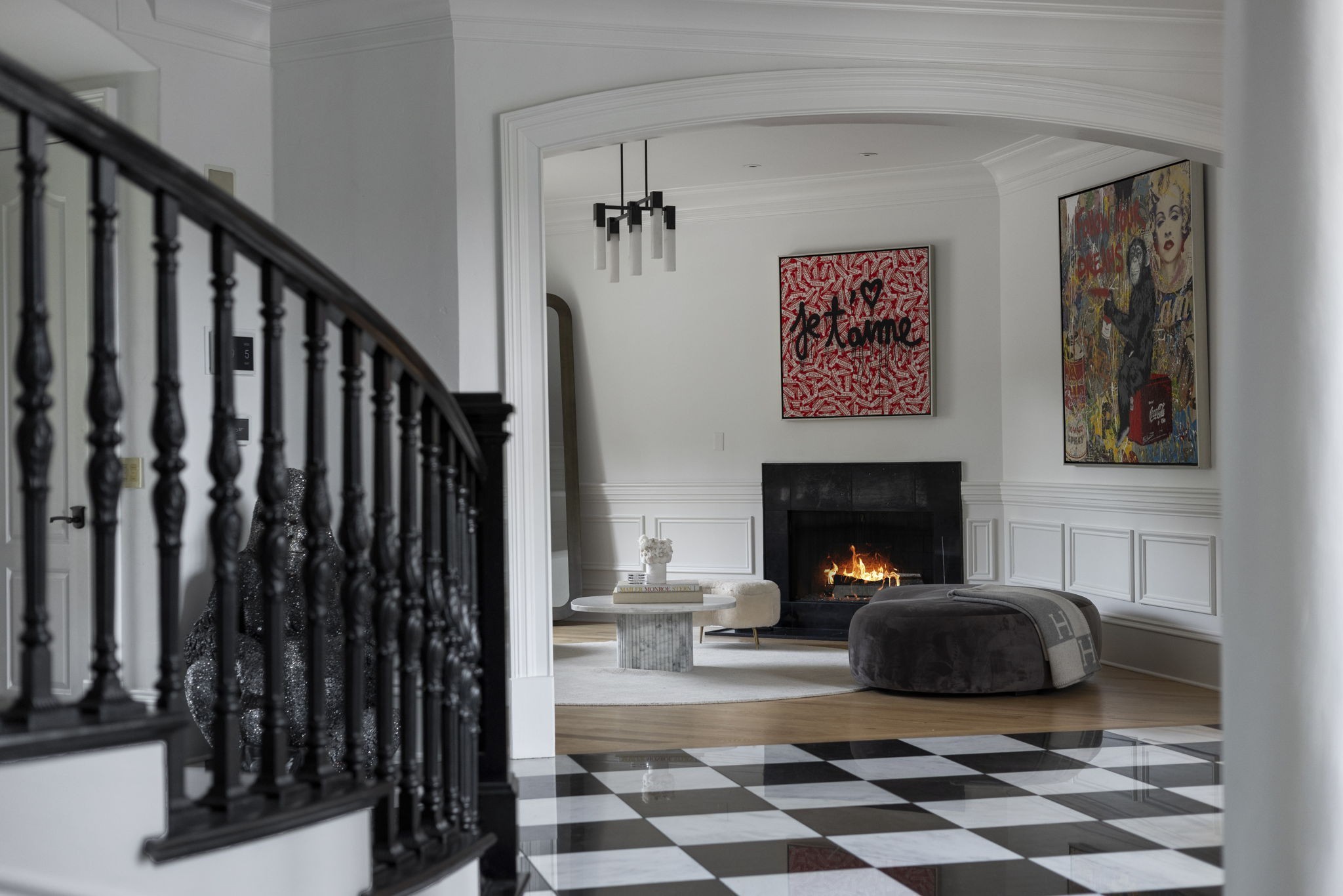 Living room with a lit fireplace, curved black staircase, colorful artwork, and checkerboard floor.