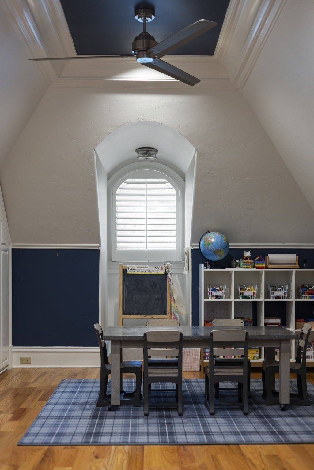Playroom with a long table, toy shelves, and an arched shuttered window beneath a vaulted ceiling.
