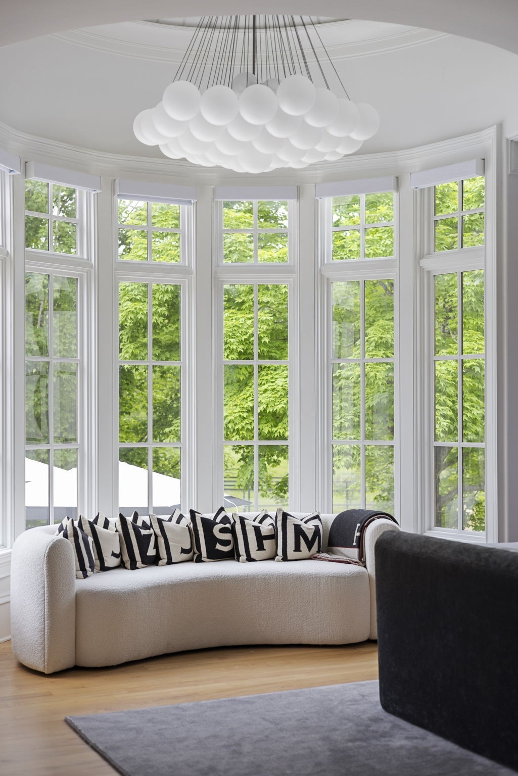 Living room with a white sofa, globe chandelier, and tall windows overlooking leafy trees.