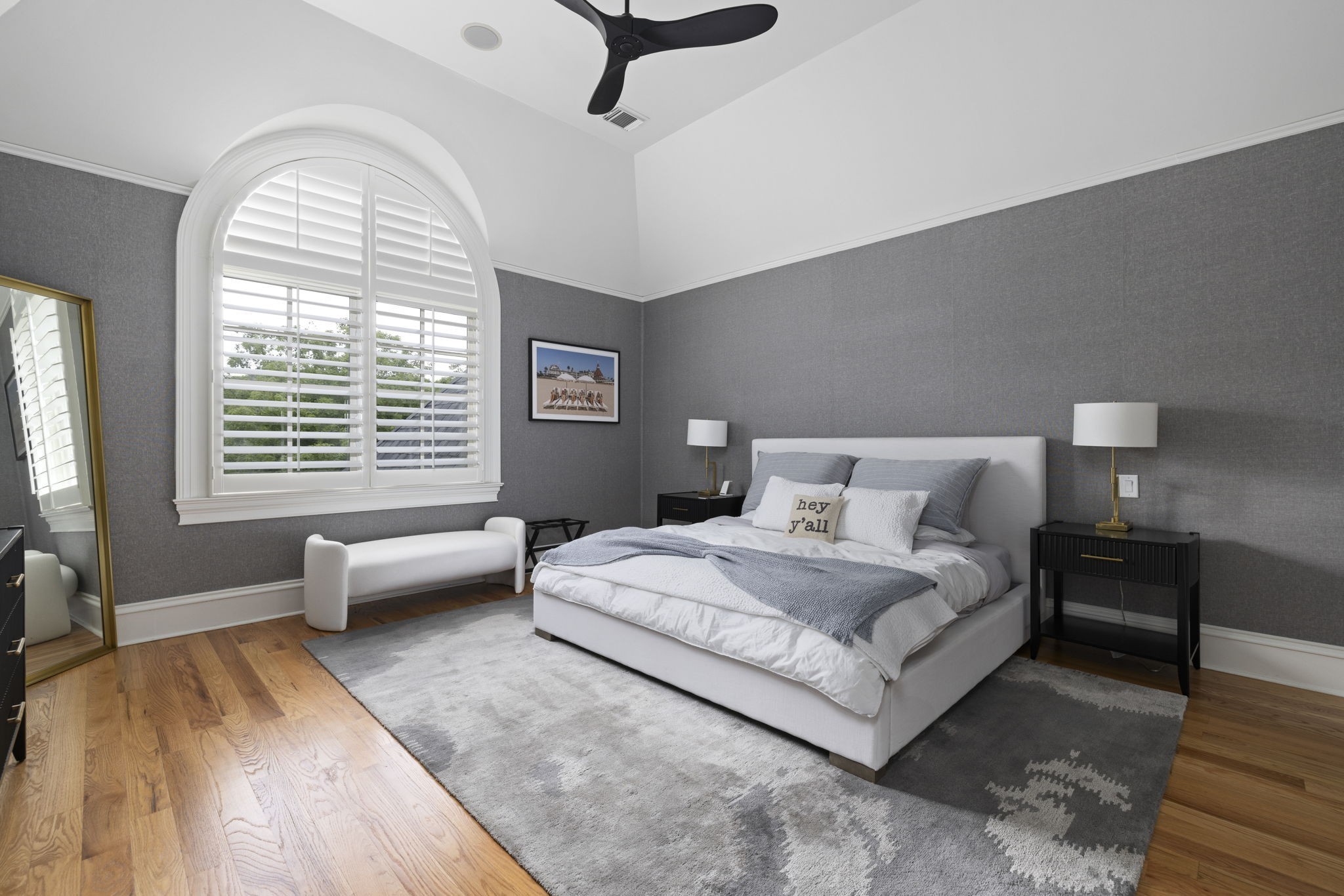 Furnished bedroom with a white bed, gray walls, and a large arched window with plantation shutters.