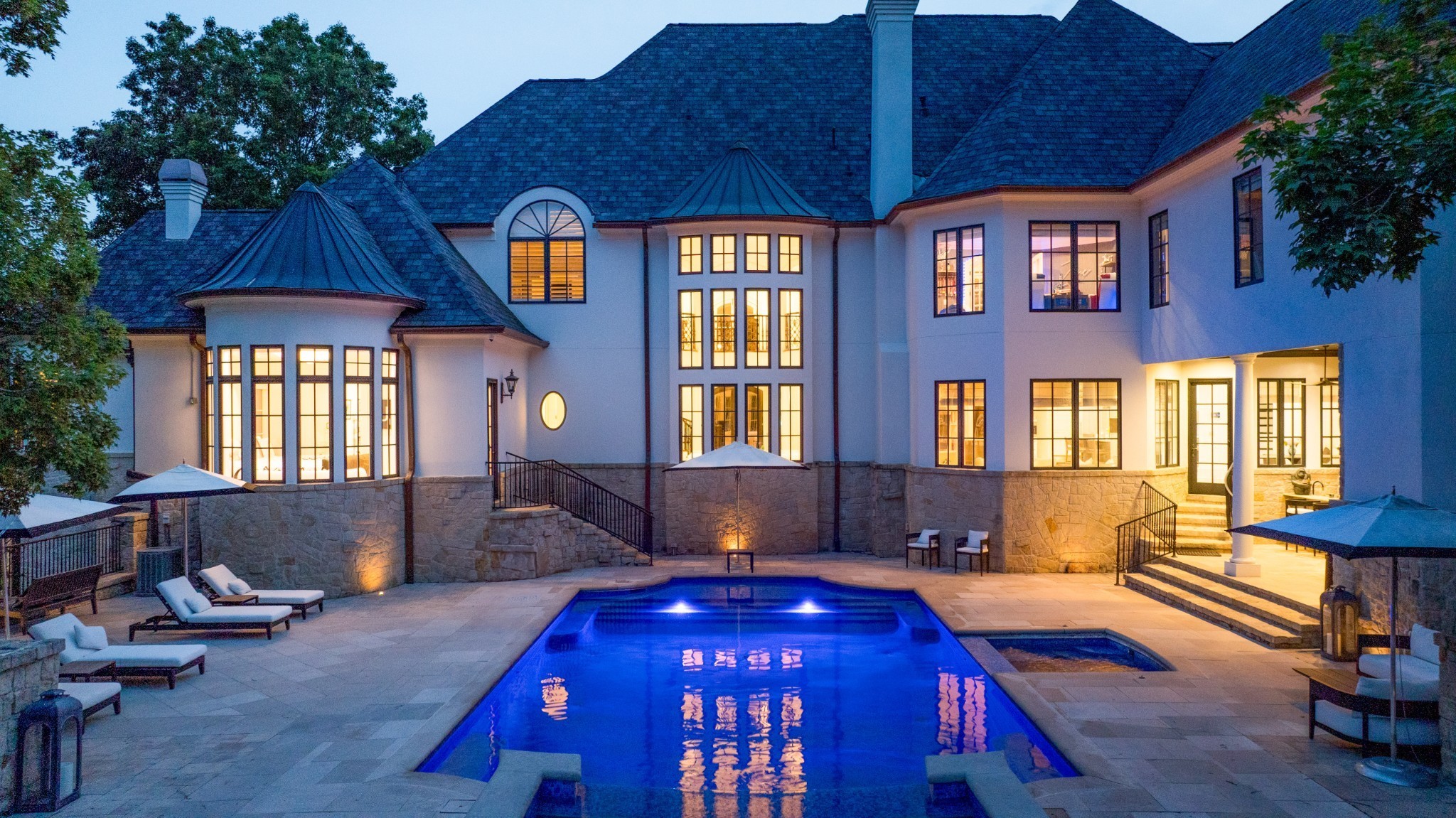 Two-story home and blue-lit swimming pool at dusk, with illuminated windows and a stone patio.
