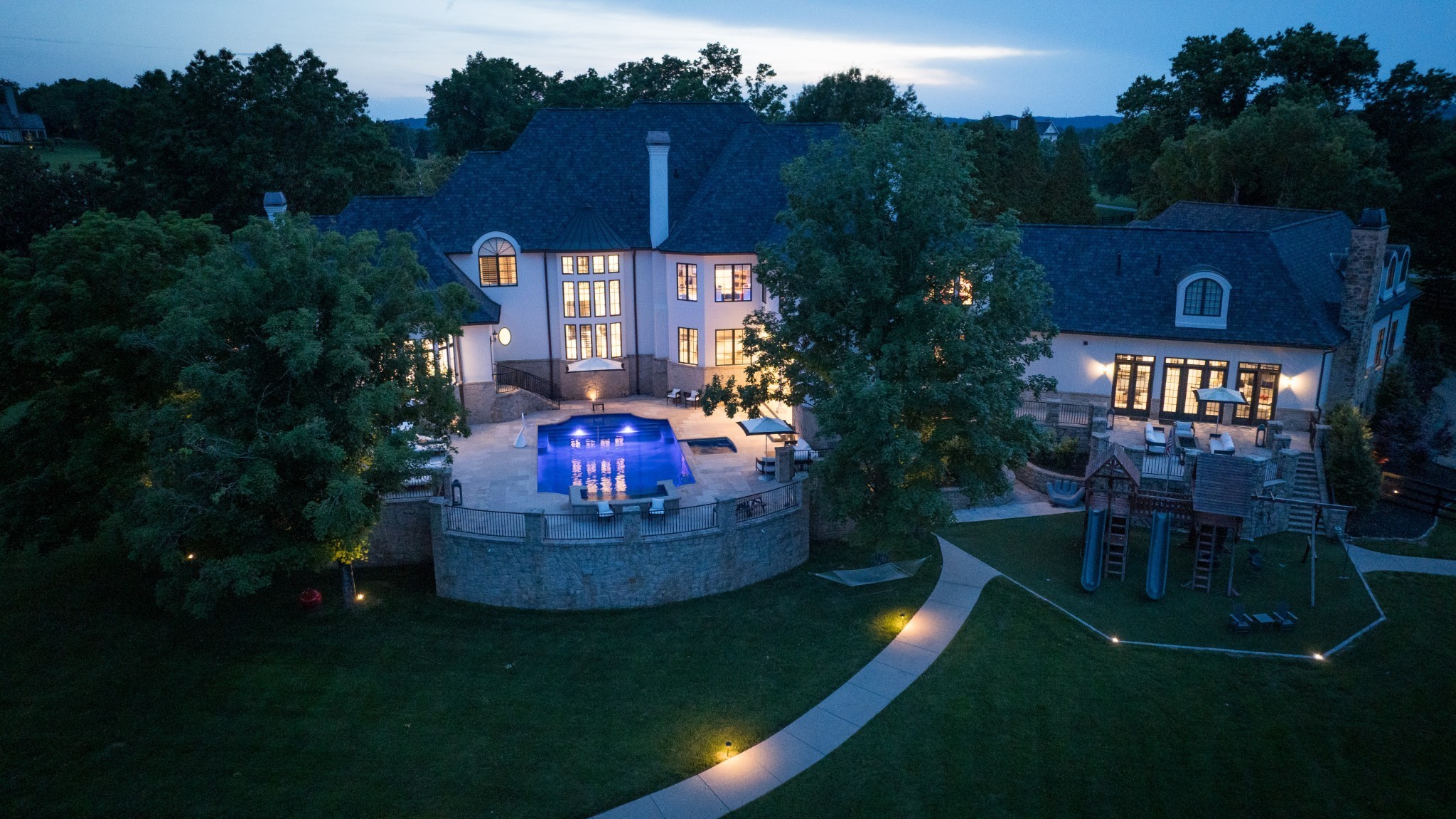 Aerial view of a large home at dusk, with a blue-lit pool, stone terrace, and expansive lawn.