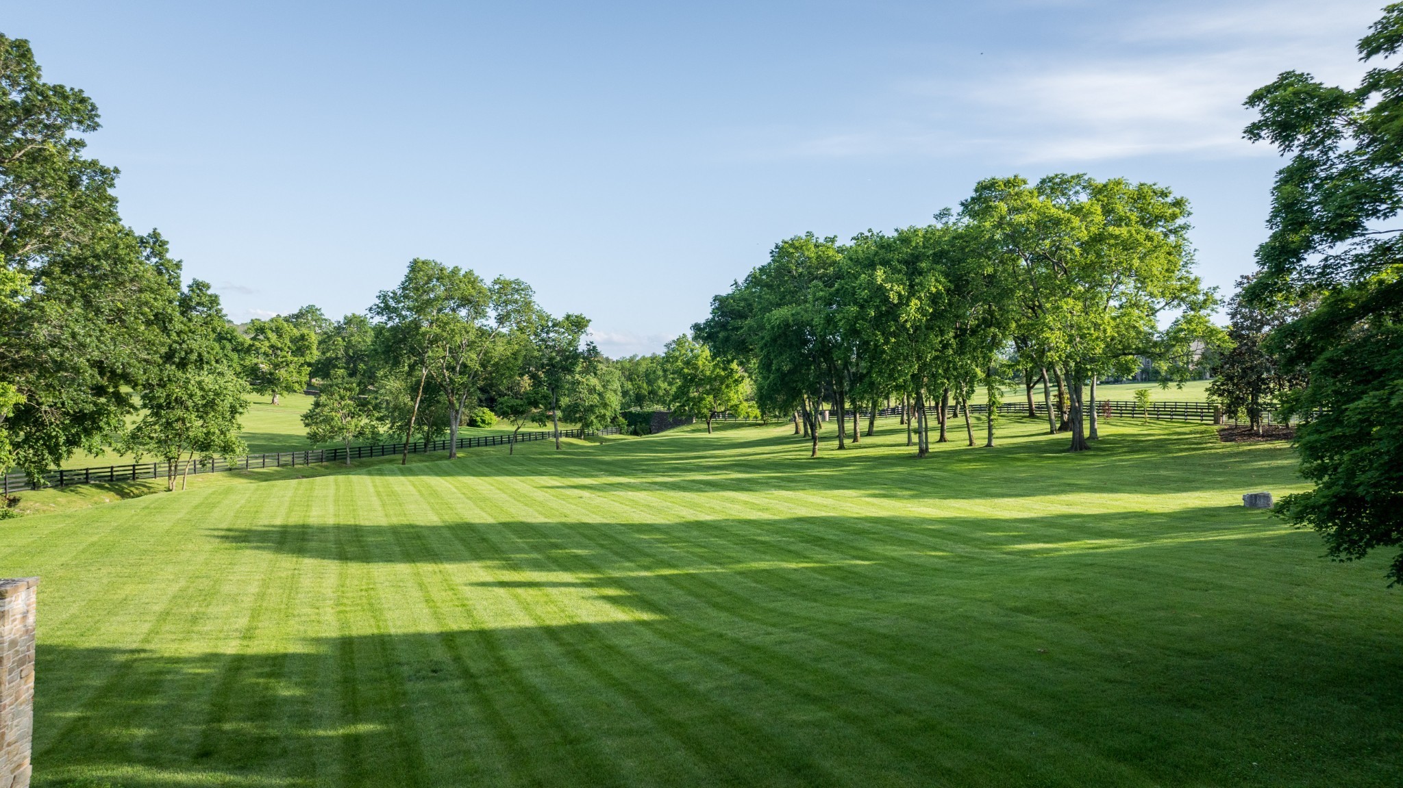 Expansive landscaped lawn with mature trees and black rail fencing.