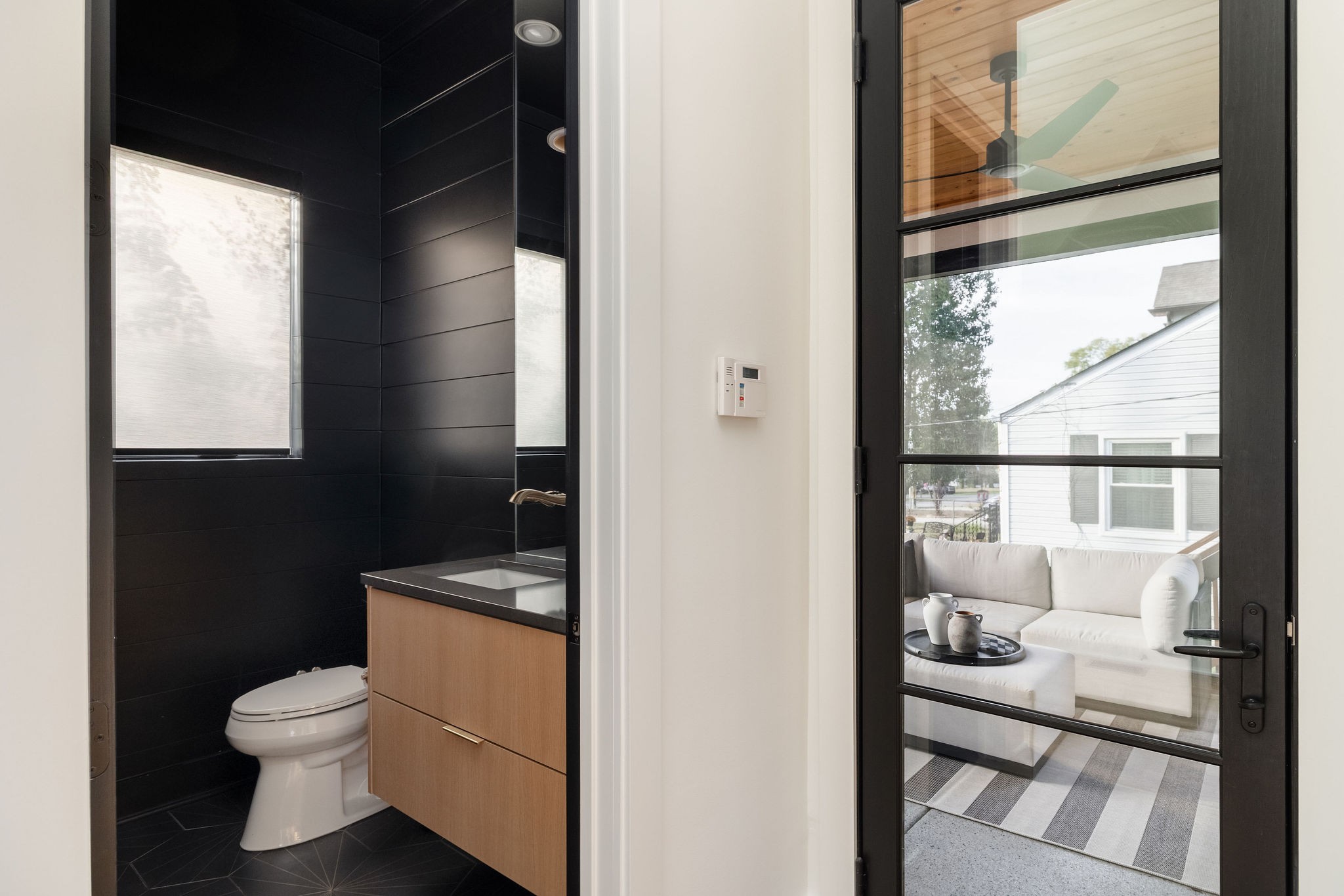 Modern black-tiled bathroom with a wood vanity beside a glass door to a furnished patio.