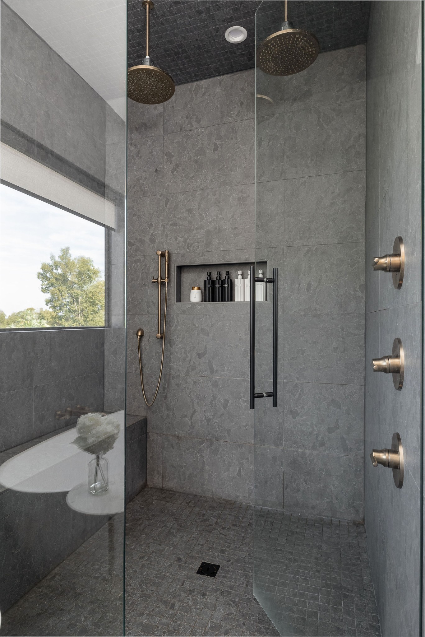 Walk-in shower with gray tile, bronze fixtures, glass enclosure, and a recessed shelf beside a window.