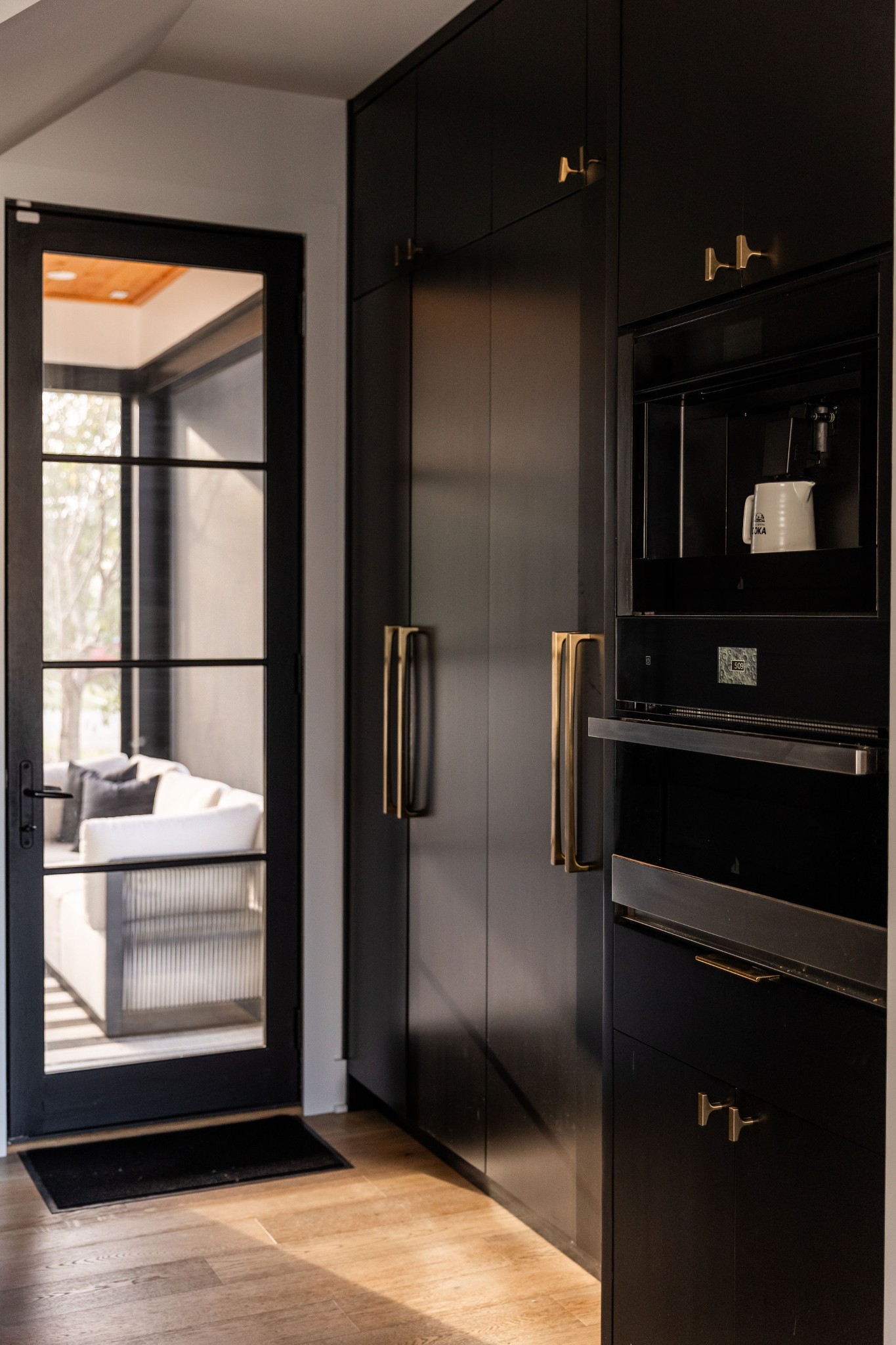 Modern kitchen with black cabinets, brass hardware, and a glass door opening to a sitting area.