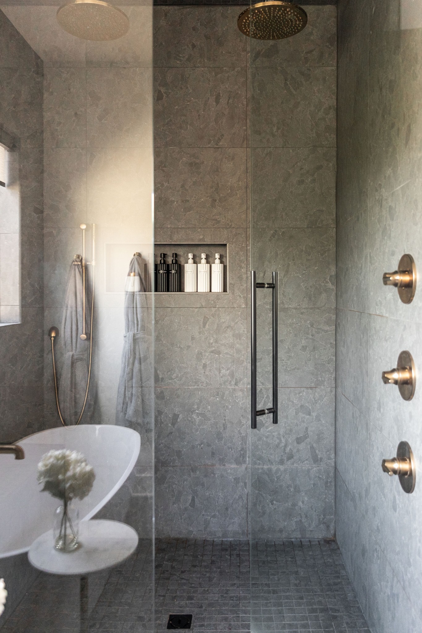 Gray tiled shower with glass enclosure, rainfall showerhead, recessed bottle niche, and brass fixtures.