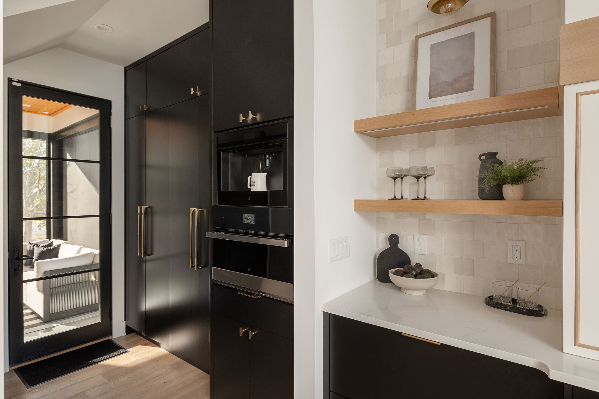 Modern kitchen with black cabinetry, brass hardware, and floating wood shelves beside a glass door.