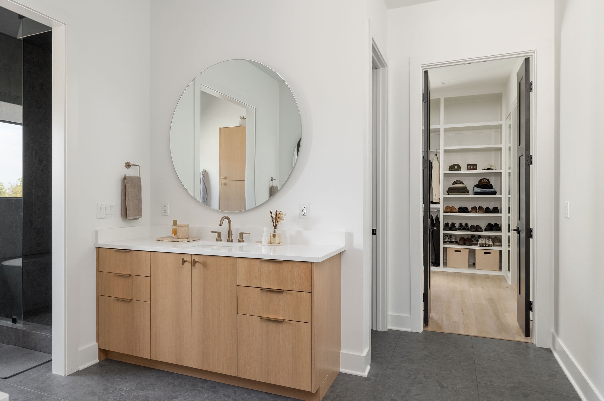 Modern bathroom vanity with a round mirror beside an open, shoe-lined walk-in closet.