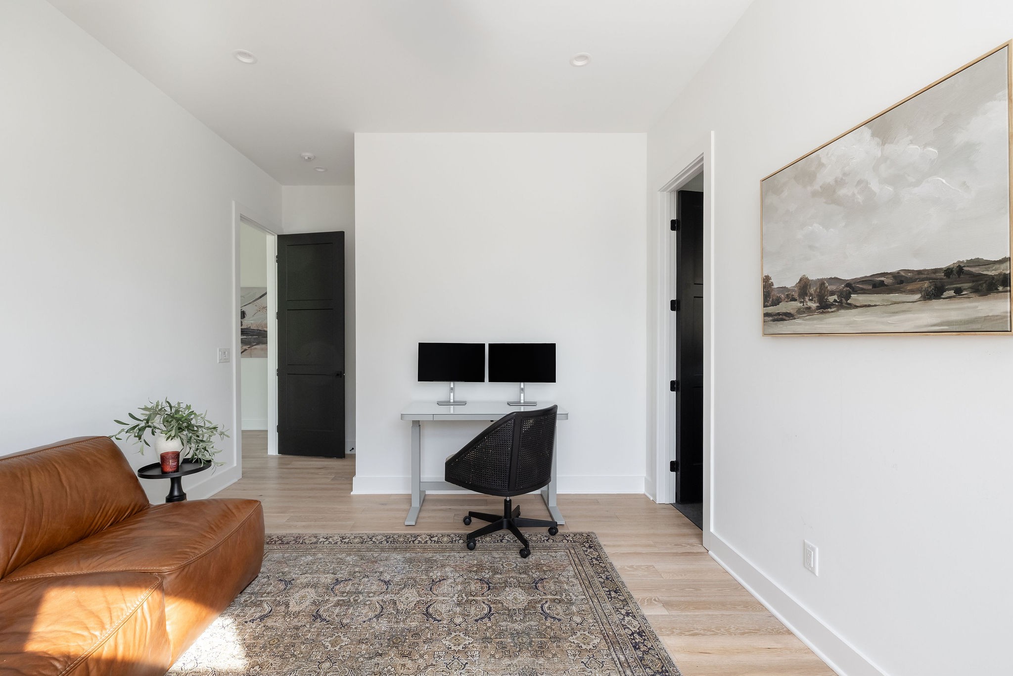 Home office with a dual-monitor desk, tan leather sofa, patterned rug, and large landscape painting.
