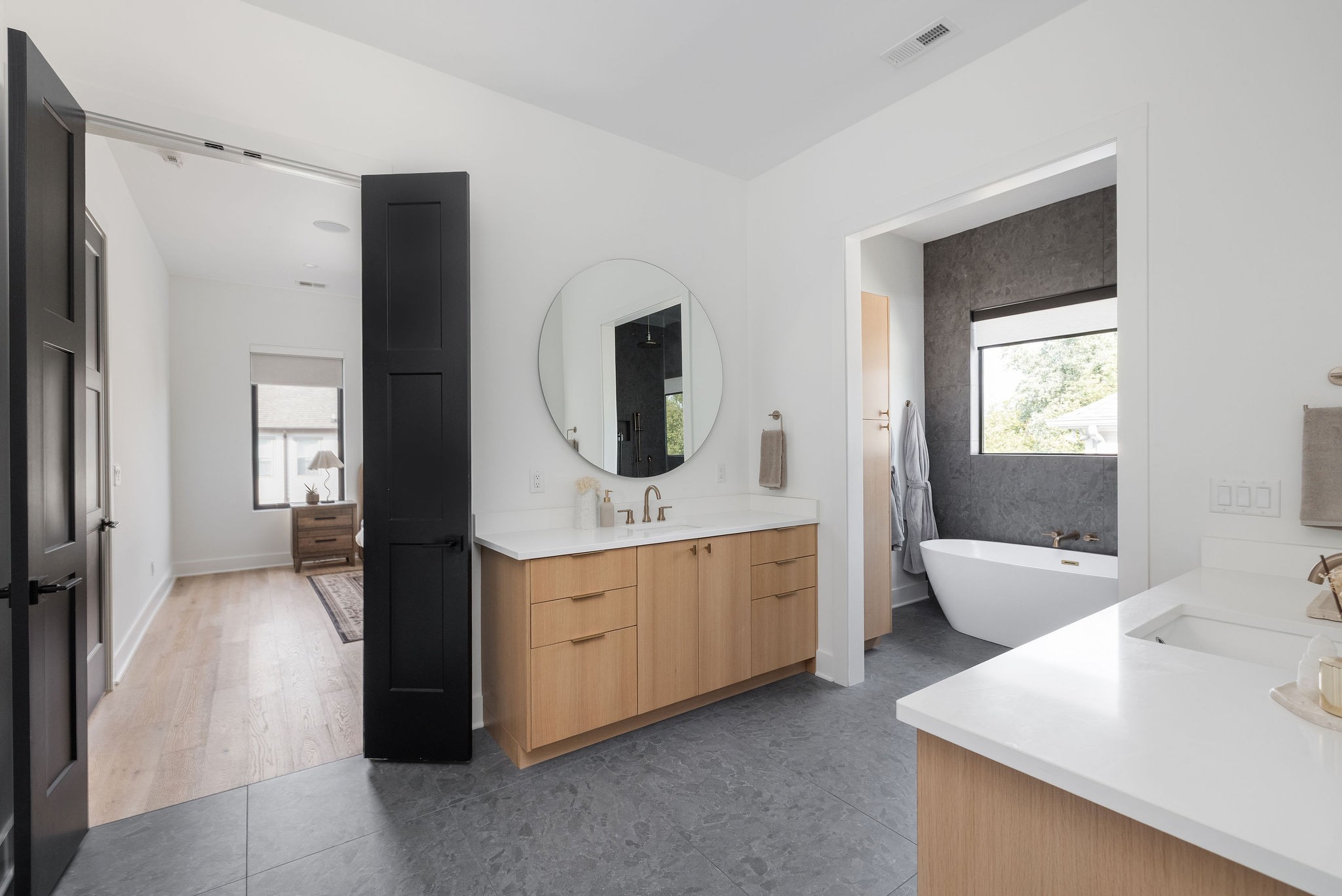 Modern bathroom with a light wood double vanity and freestanding tub against a charcoal tile wall.
