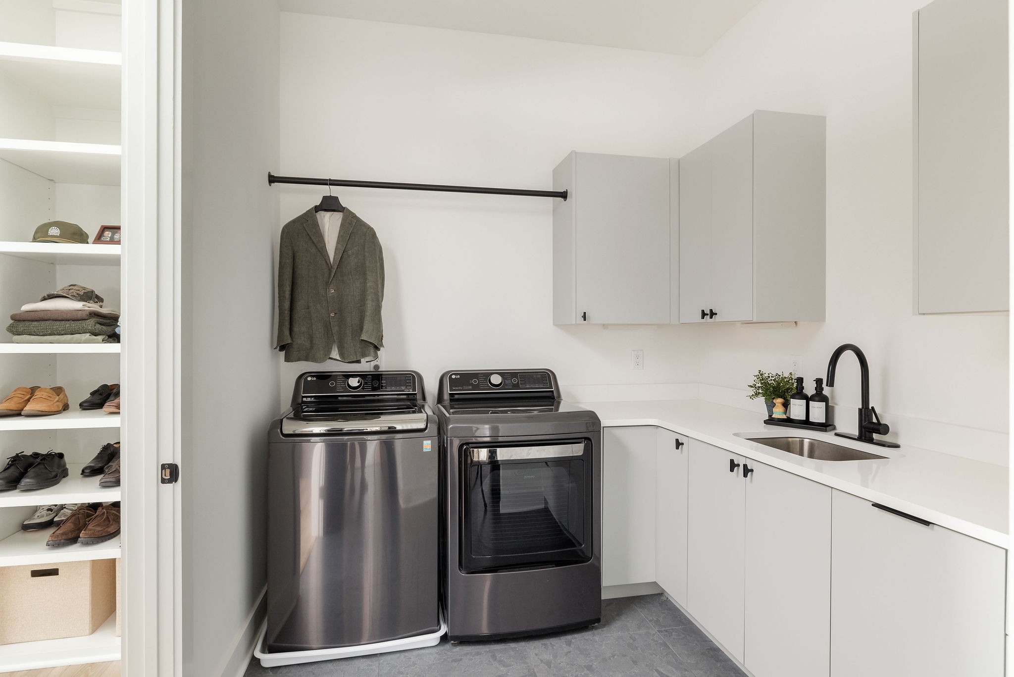 Laundry room with gray washer and dryer, white cabinetry, sink, and open shoe shelves.