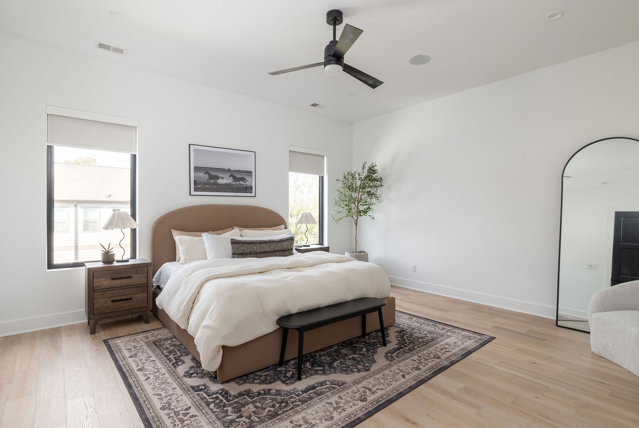 Bright bedroom with a king-size bed, rounded tan headboard, patterned rug, and tall arched mirror.