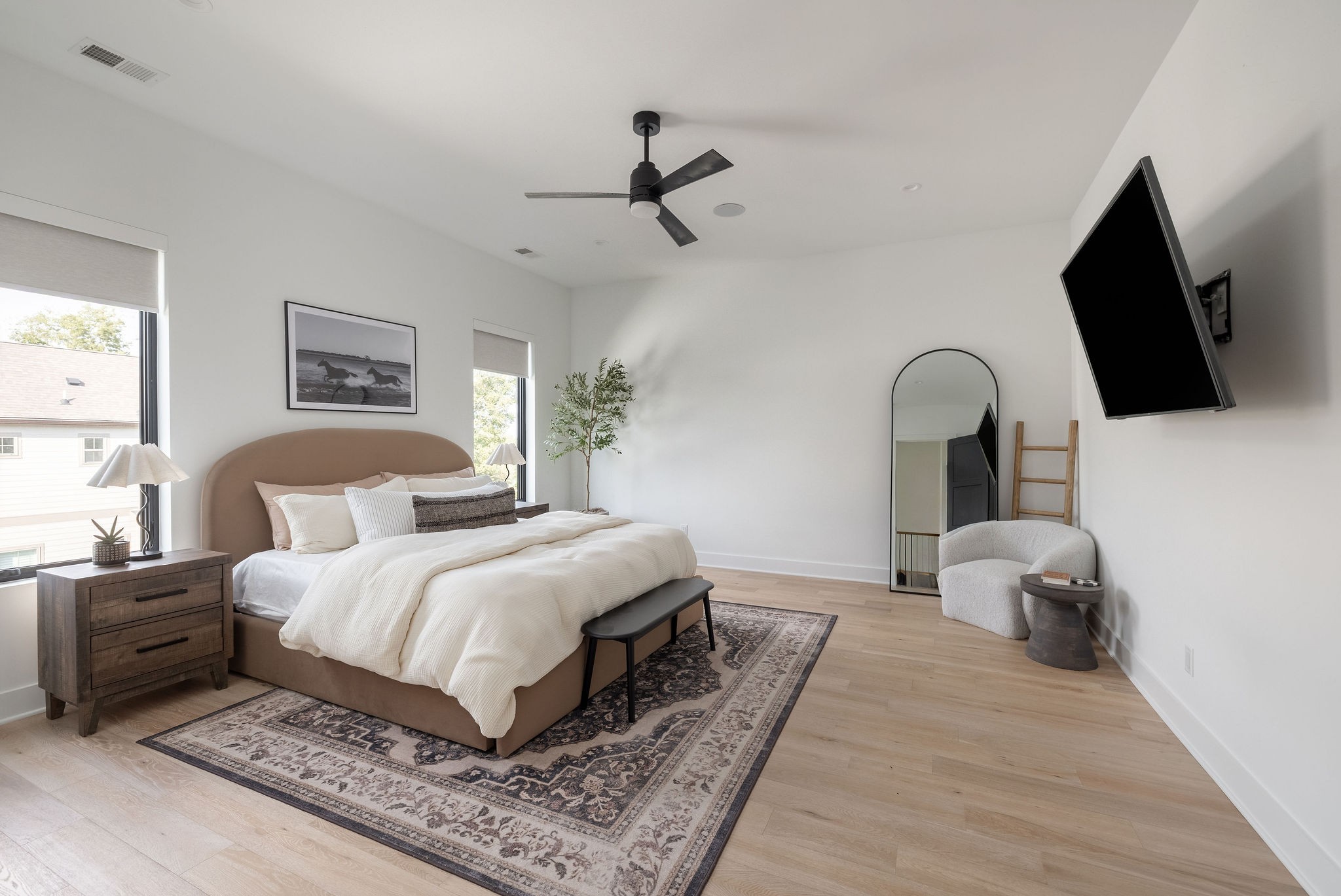 Staged bedroom with a tan upholstered bed, patterned rug, arched mirror, and wall-mounted television.