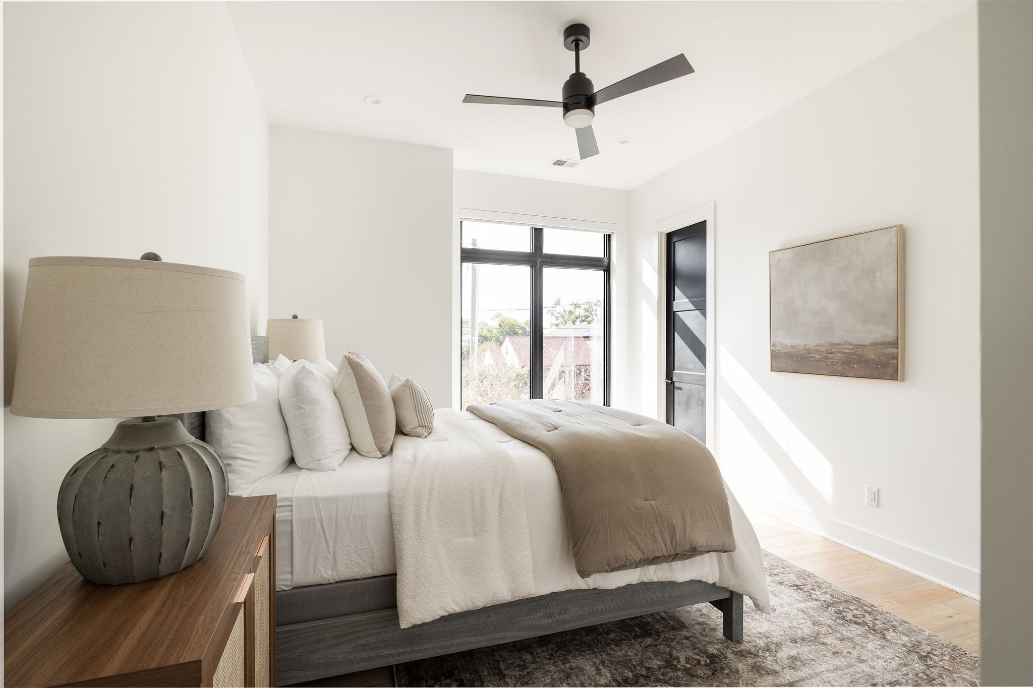 Bright staged bedroom with a white-and-taupe bed, black-framed window, and dark ceiling fan.
