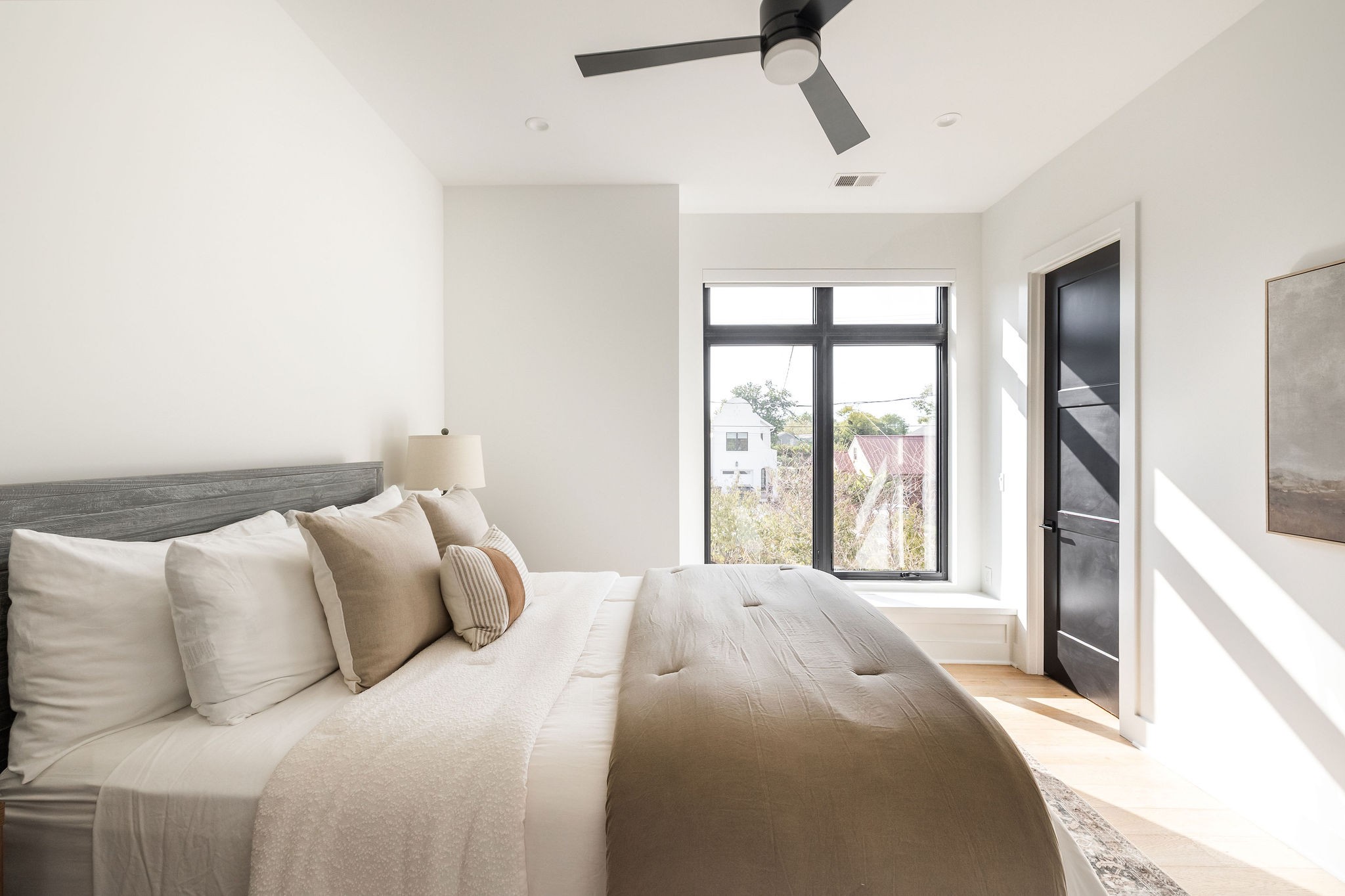 Bright bedroom with a taupe-and-white bed, black-framed windows, and a dark ceiling fan.