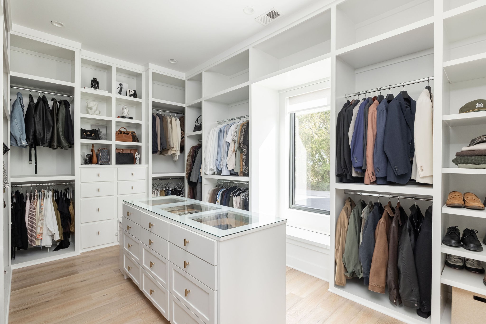 Walk-in closet with a glass-topped island, extensive white shelving, and a large window.