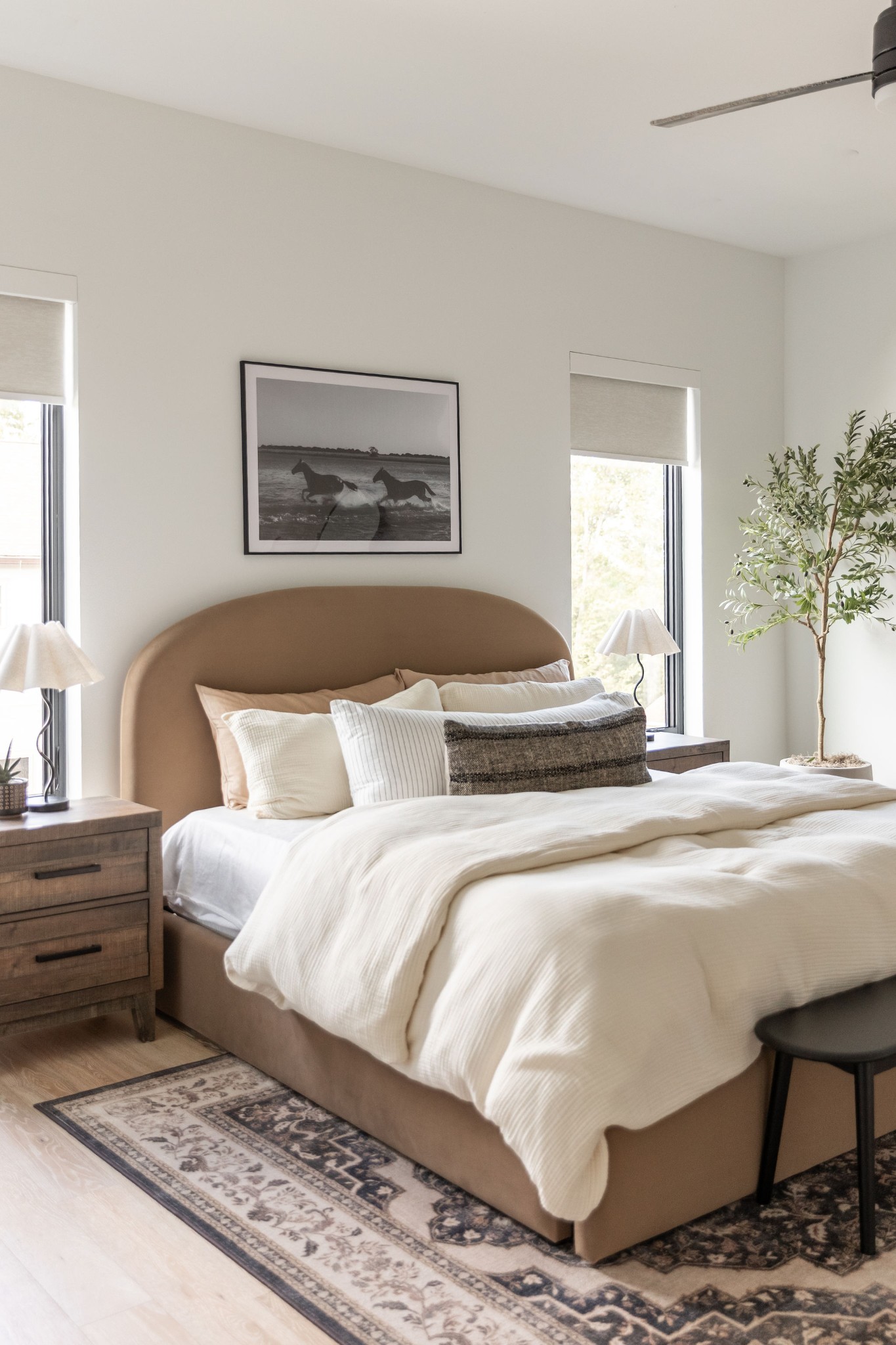Staged bedroom with a curved tan headboard, white bedding, tall windows, and a patterned rug.
