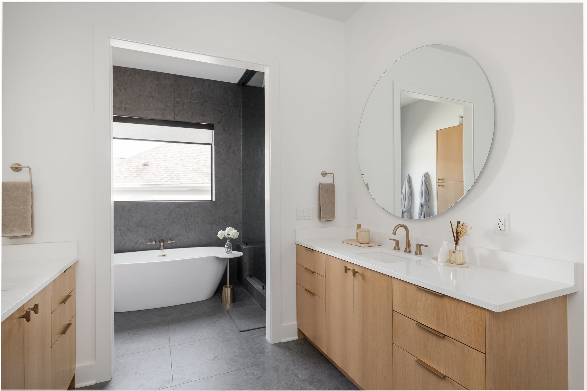 Modern bathroom with a wood double vanity and round mirror beside a freestanding tub in a dark tiled alcove.
