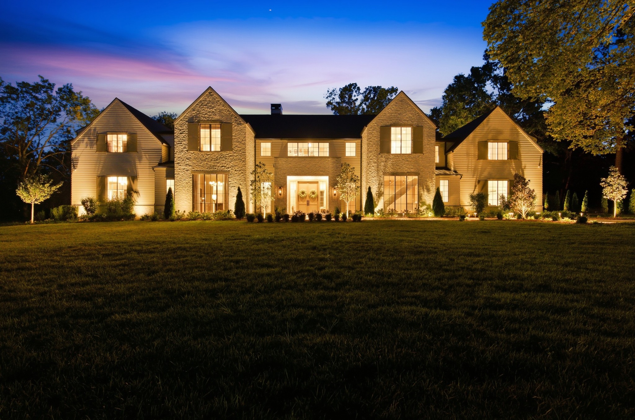 Large stone-and-siding home with illuminated windows and a broad front lawn at dusk