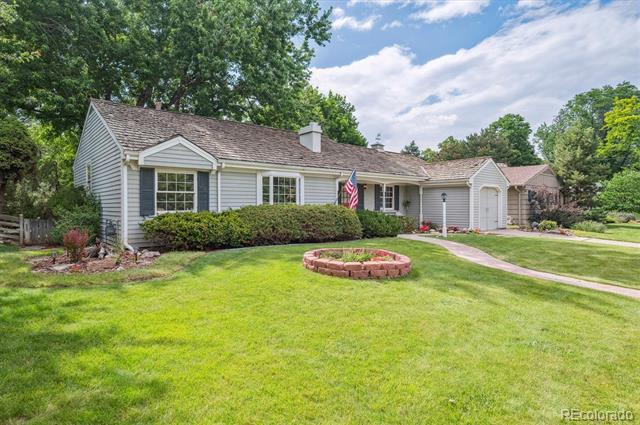 Single-story gray house with an attached garage, broad lawn, circular brick planter, and American flag by the entrance.
