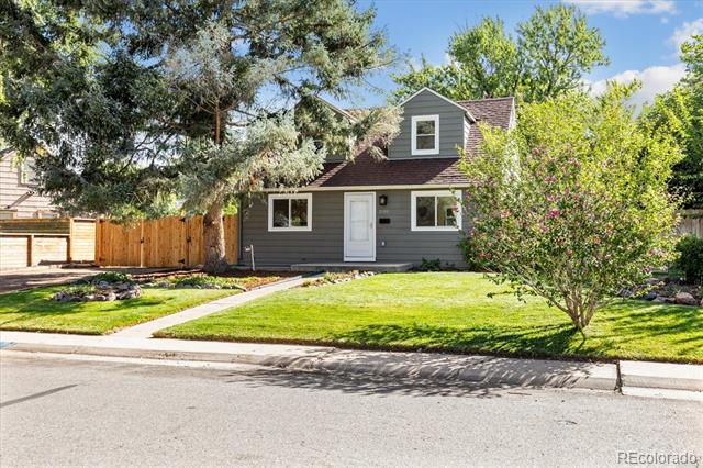 Gray house with a small dormer, white trim, green lawn, mature trees, and flowering shrubs.