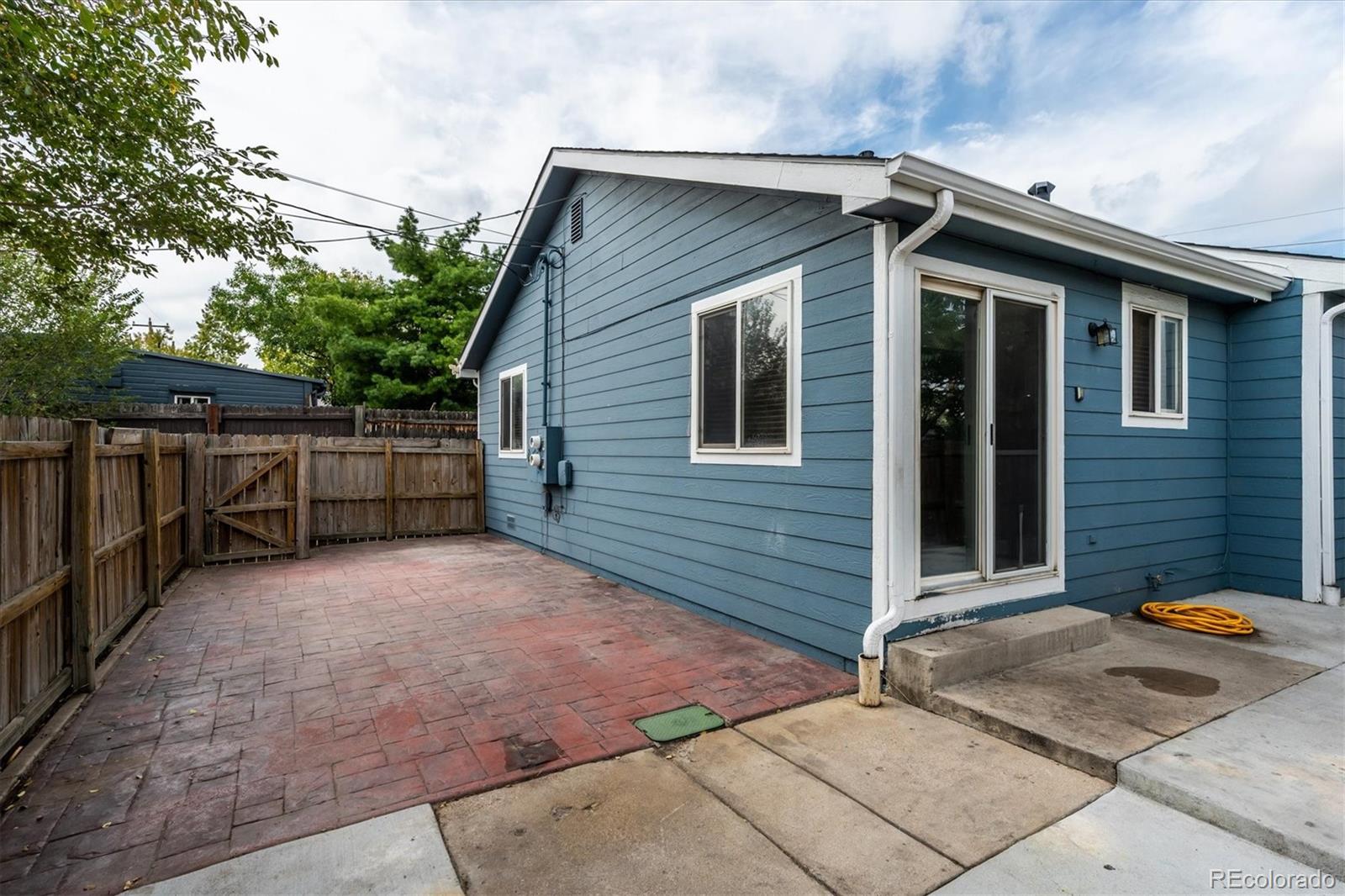Blue-sided house beside a fenced red-brick patio and concrete walkway.