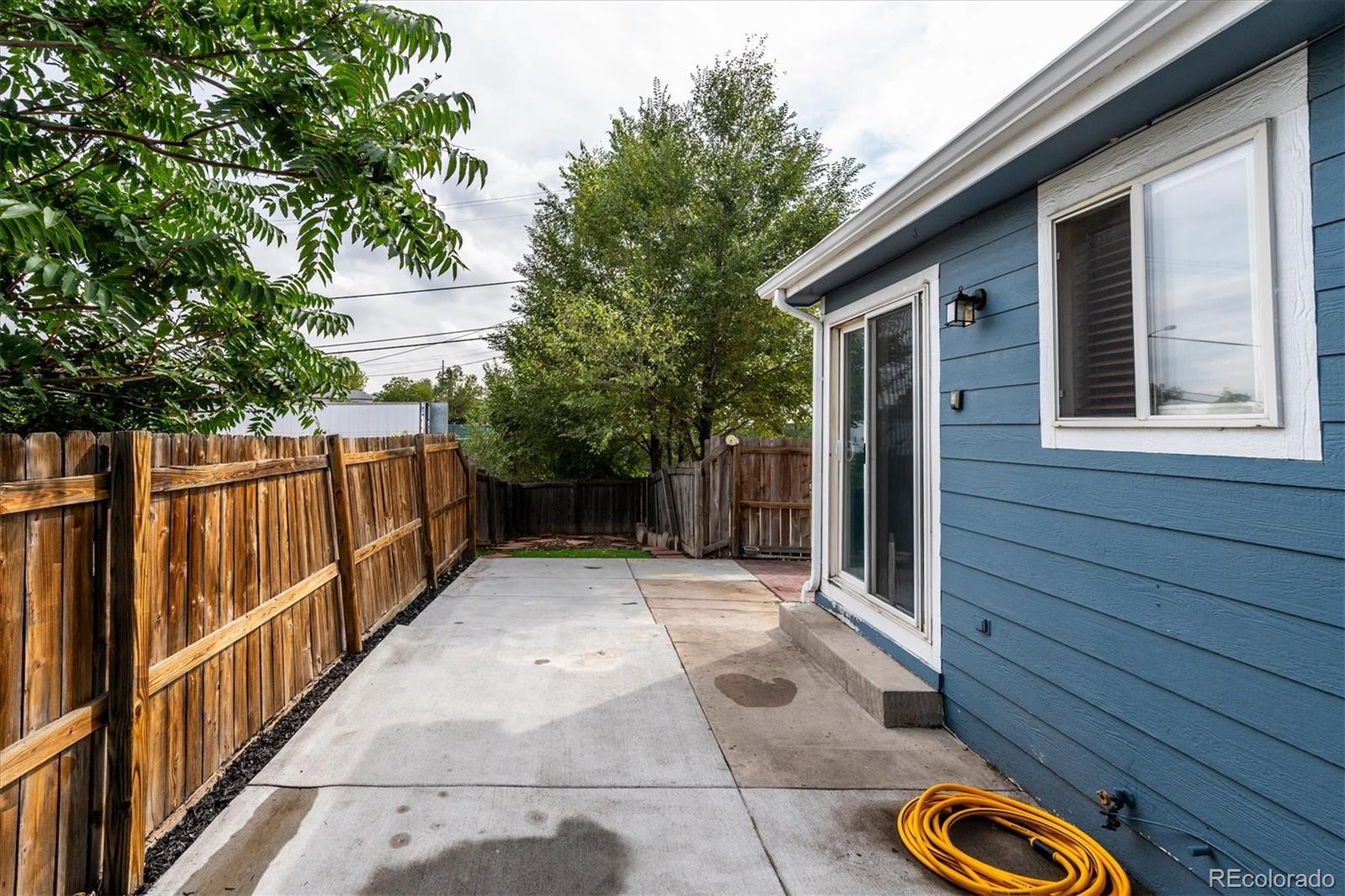Concrete patio beside a blue house, enclosed by a wooden fence with trees at the far end.