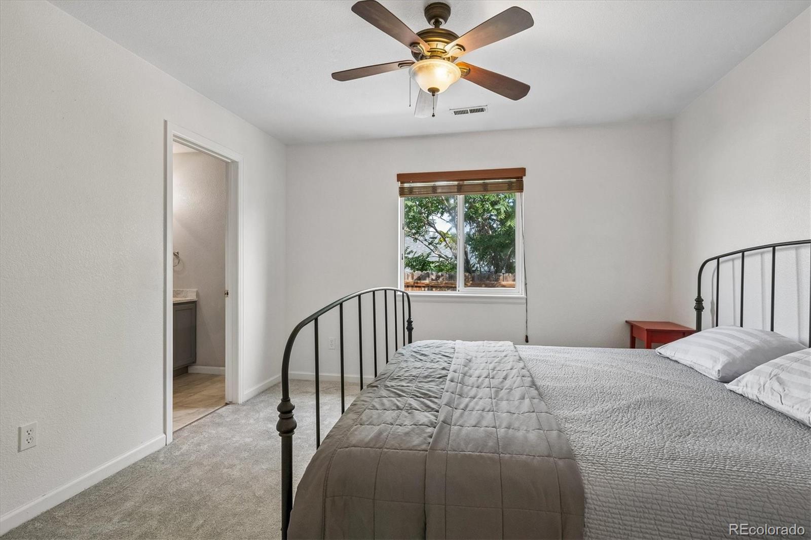 Bedroom with a black metal bed frame, gray bedding, ceiling fan, and window overlooking greenery.