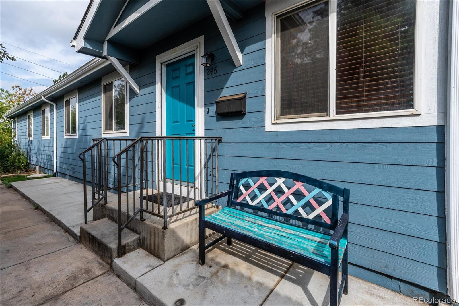 Blue-sided home with a turquoise front door, concrete entry steps, black railings, and a colorful bench.
