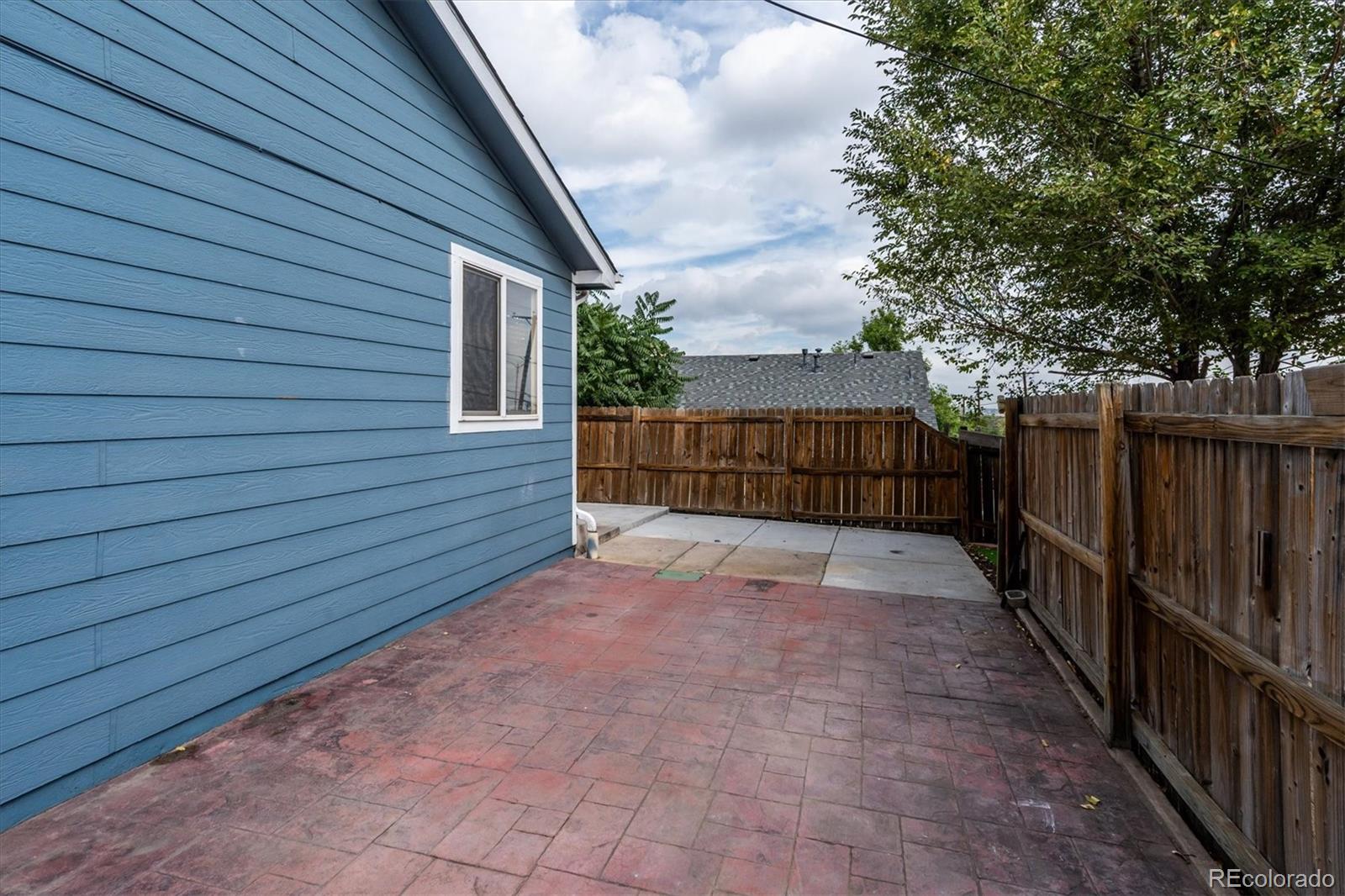 Paved backyard patio beside a blue-sided house, enclosed by wooden fences beneath a leafy tree.
