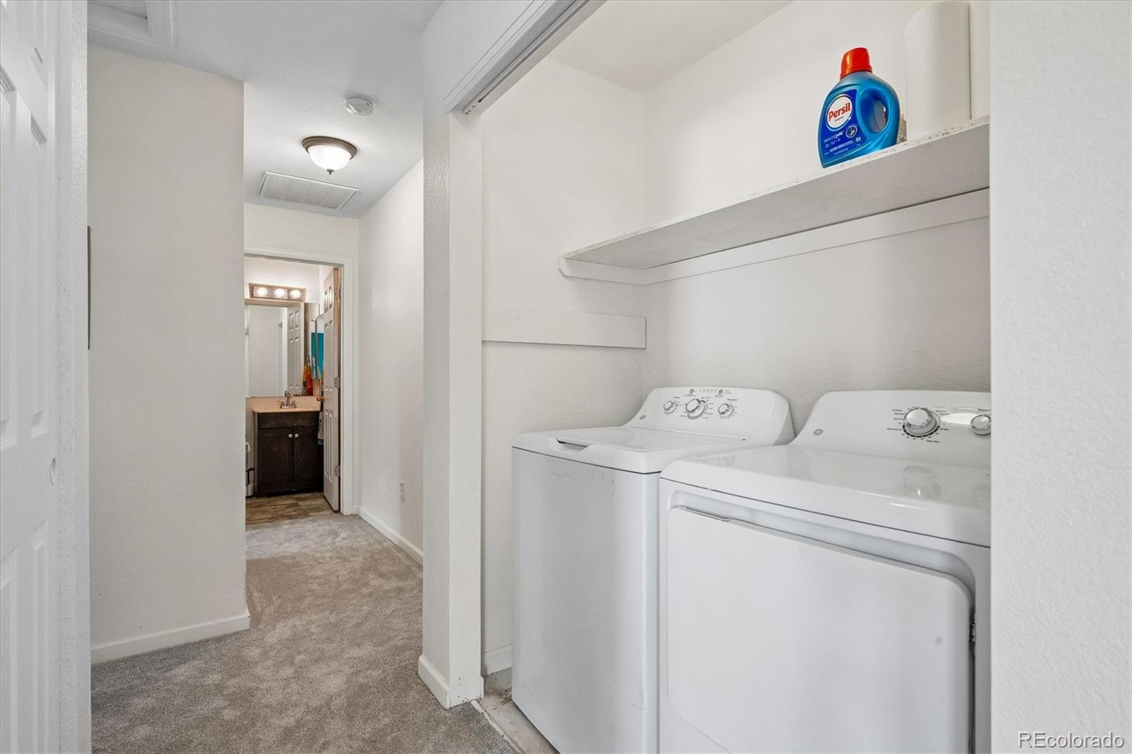 Laundry room with white washer and dryer, a detergent bottle on the shelf, and a bathroom vanity beyond.