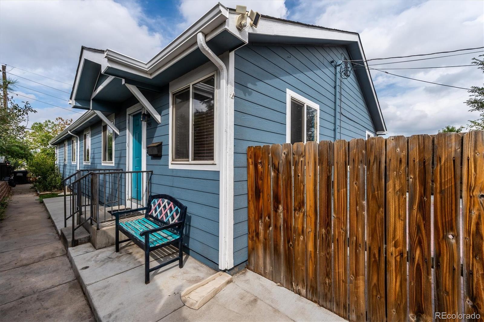 Blue house with a turquoise front door, concrete entry patio, wooden privacy fence, and outdoor bench.