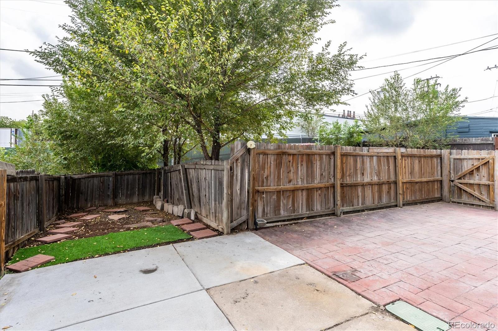 Backyard with a brick patio, tall wooden fence, mature tree, and small lawn.