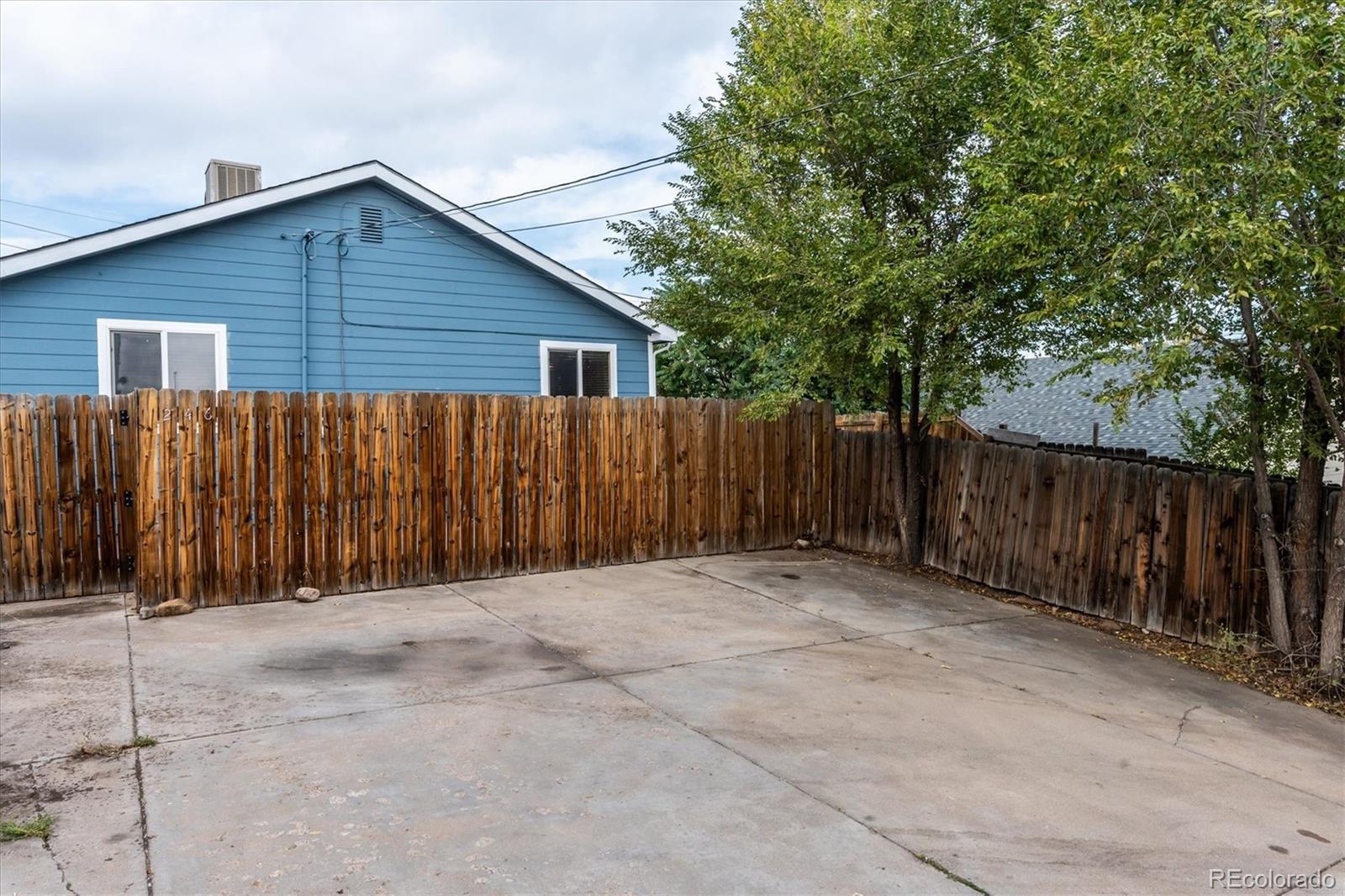 Concrete backyard patio enclosed by a wooden privacy fence, with leafy trees and a blue house beyond.