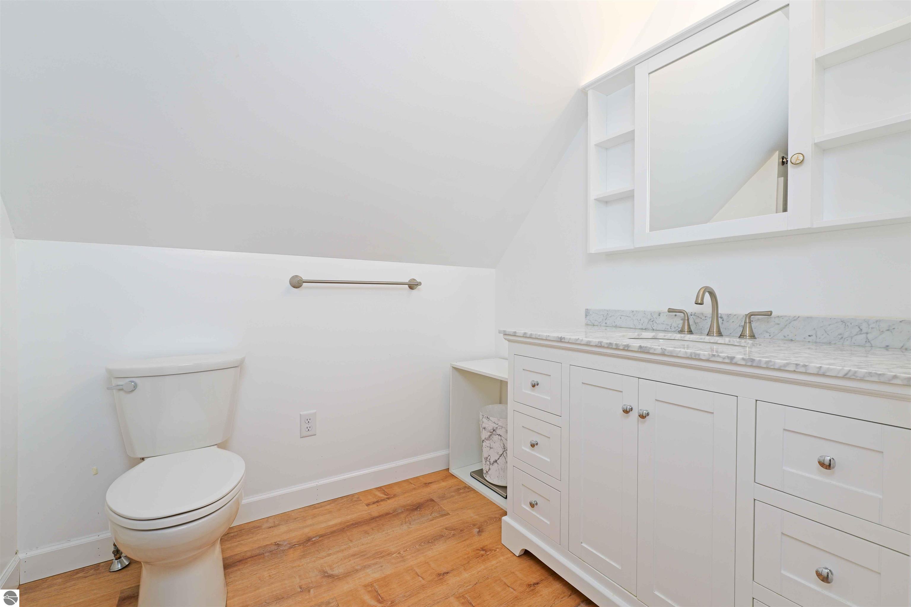 Bathroom with a marble-topped white vanity, brass fixtures, toilet, and warm wood flooring.