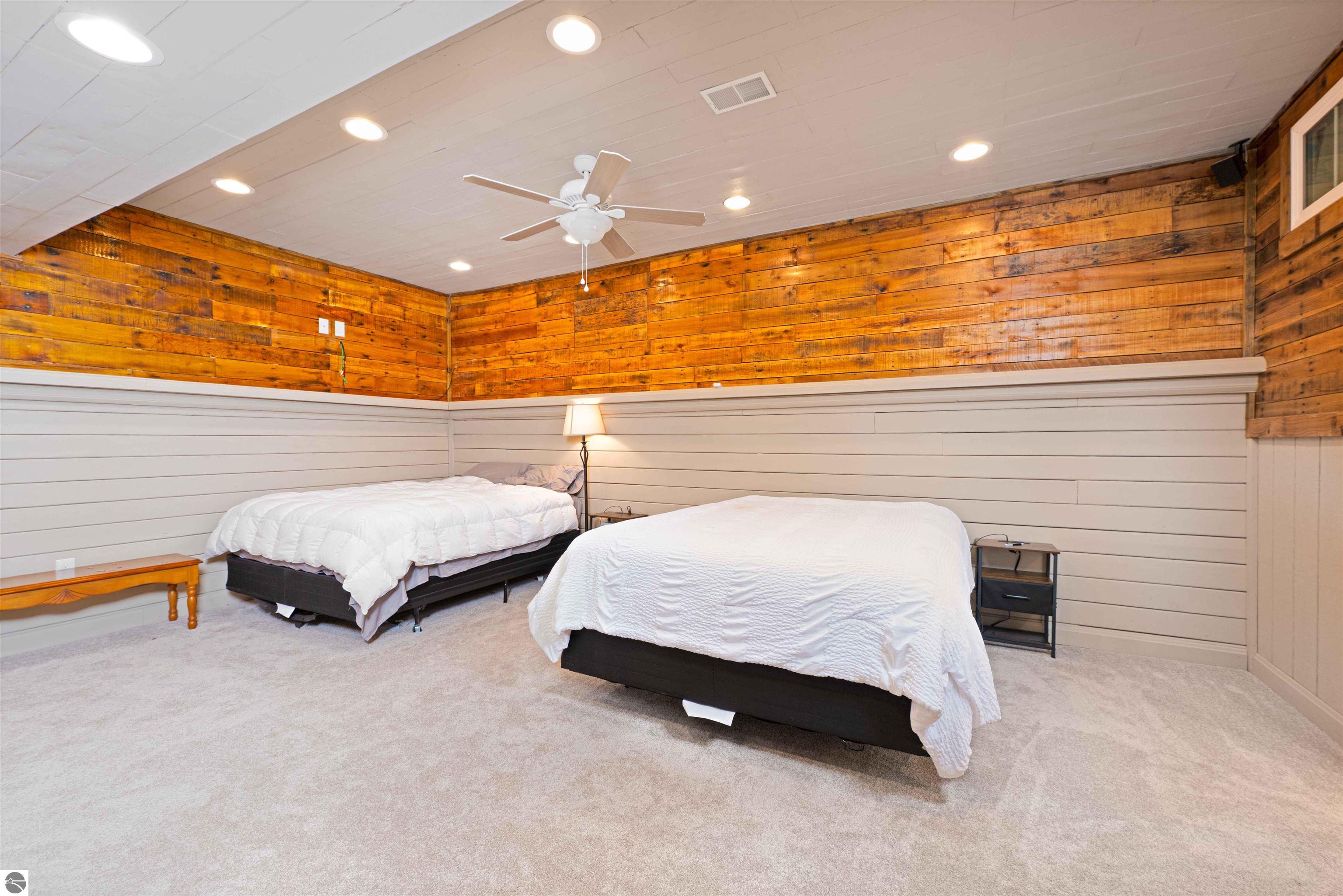 Bedroom with two white-covered beds, warm wood plank walls, beige carpet, and a ceiling fan.