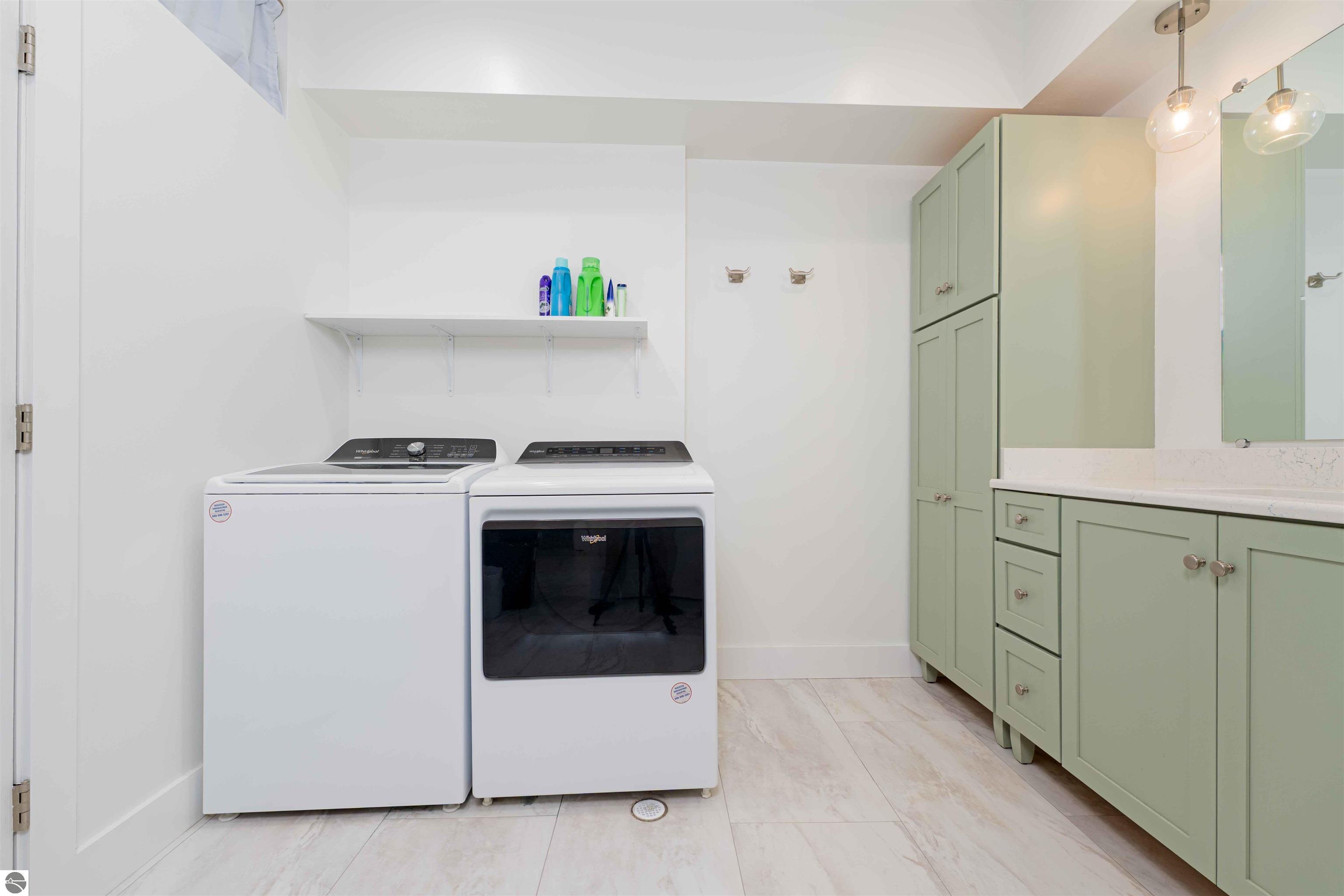 Laundry room with white washer and dryer beside sage-green cabinets and a pale stone countertop.