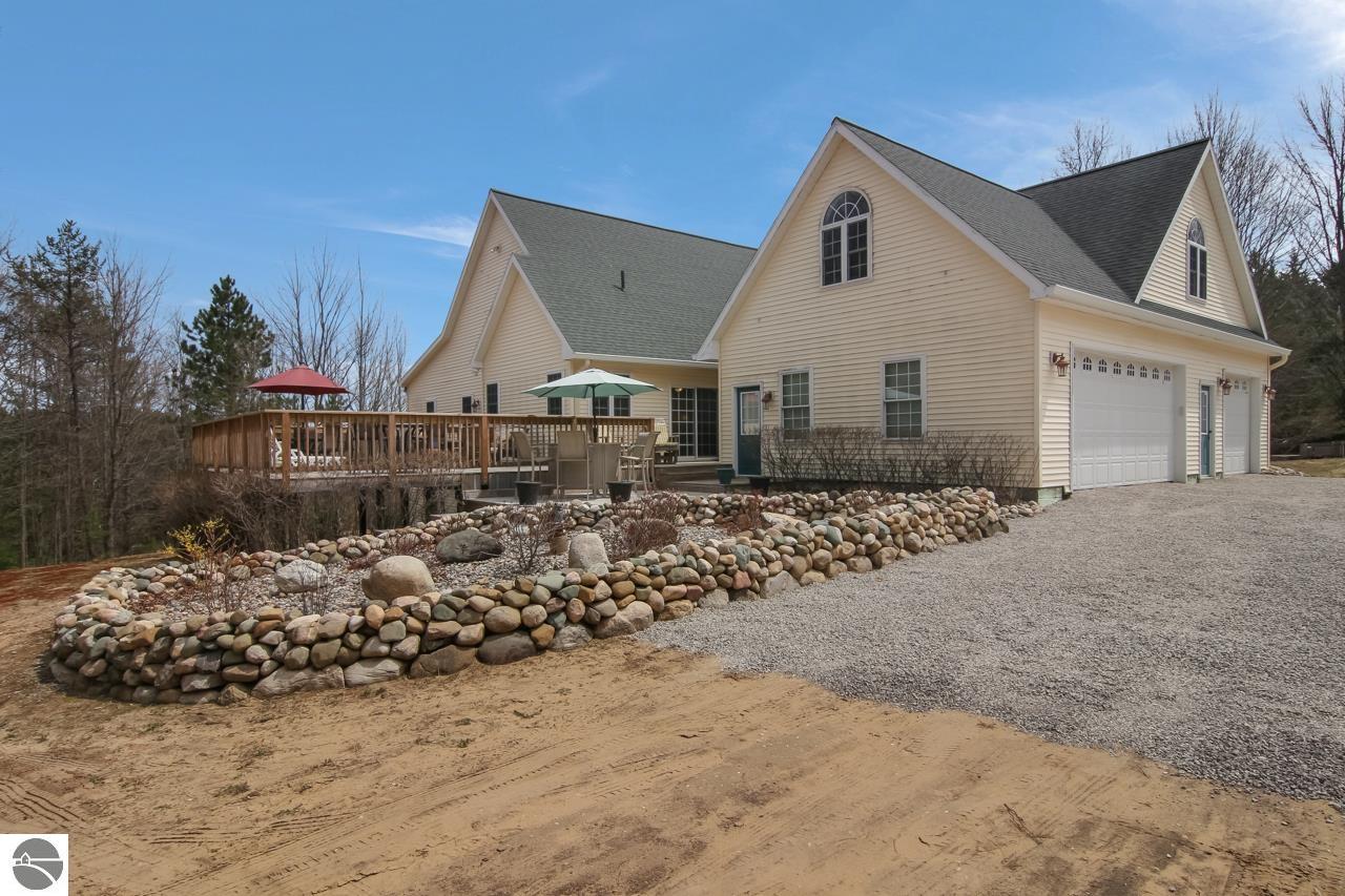 Beige home with a raised deck, stone retaining wall, and broad gravel driveway.