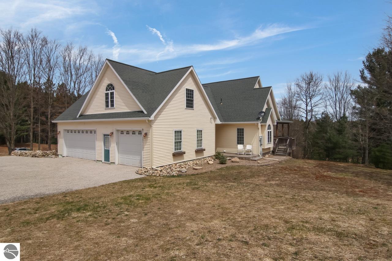 Beige gabled home with an attached two-car garage, gravel driveway, and wooded surroundings.